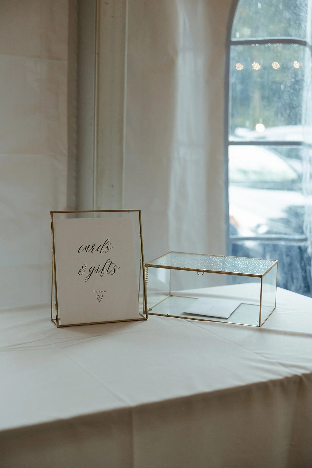 A sign reading 'cards & gifts' next to a glass display box on a table with a white tablecloth, near a window with curtains letting in natural light.