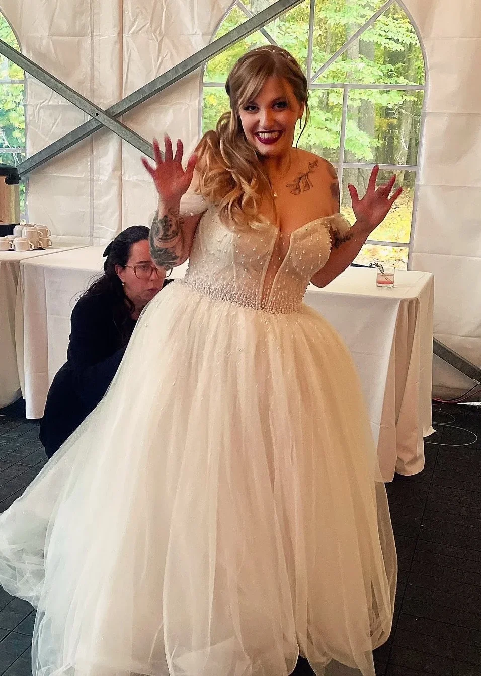 Woman in an off-the-shoulder, cream-colored ballgown with tattoos on her arms and chest, smiling and raising her hands, standing inside a tent with greenery visible outside.