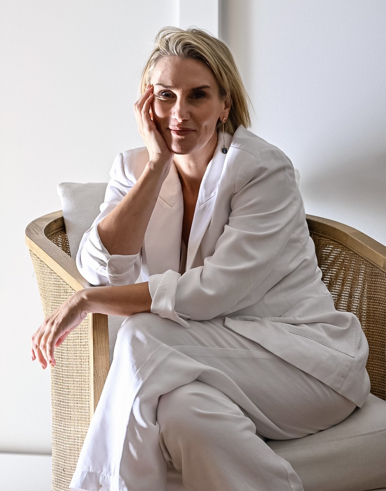 A woman with blonde hair sitting on a wooden chair with a woven backrest, wearing a white outfit, resting her head on her hand, looking at the camera with a slight smile.
