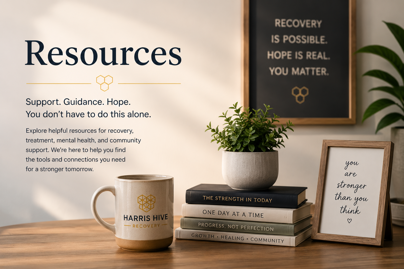 A desk with a sign titled 'Resources,' a mug with 'Harris Hive Recovery' on it, a stack of books with titles about strength and growth, a framed quote reading 'you are stronger than you think,' and a potted plant. In the background, there is a black and white motivational sign on the wall.