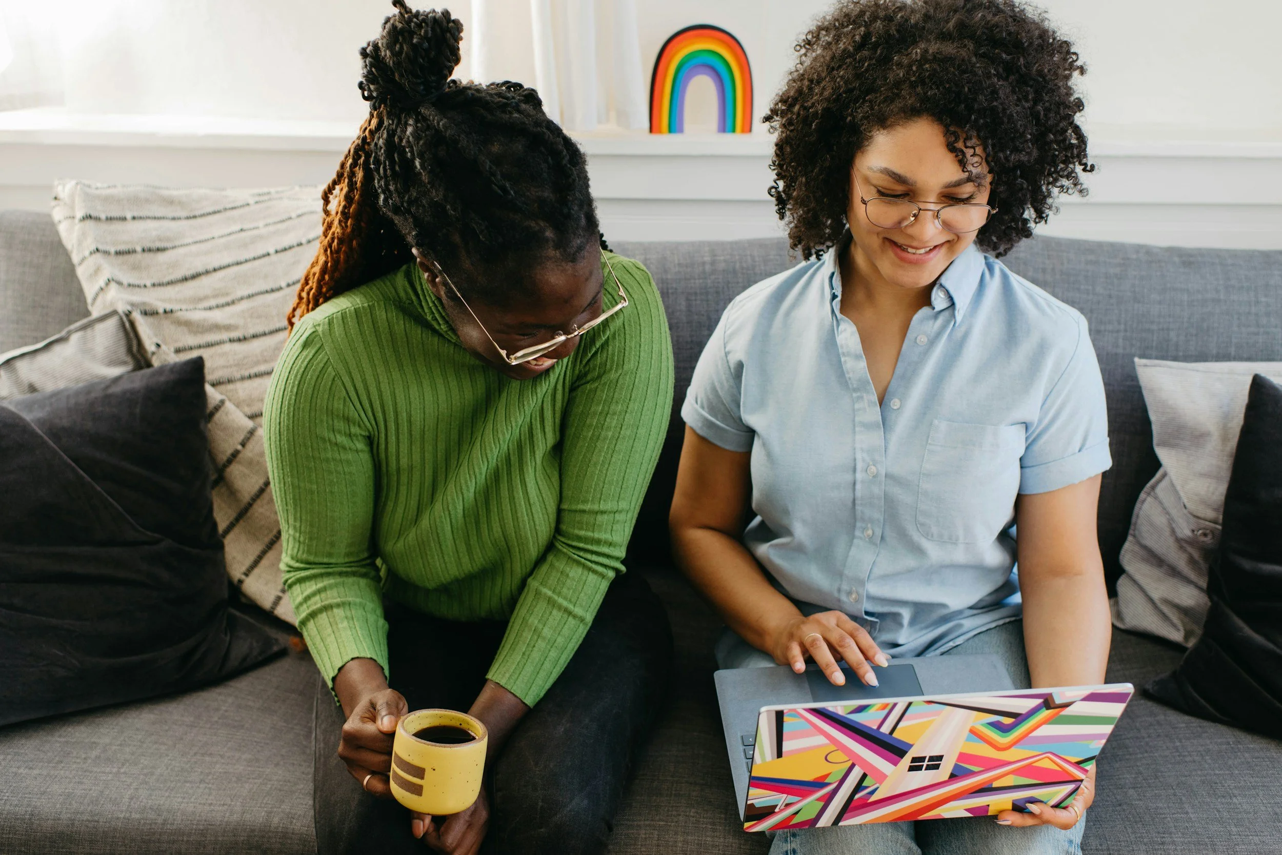 Two women sitting on a gray sofa, looking at a colorful laptop. One woman, wearing glasses and a green sweater, is holding a cup of coffee, and the other woman, with curly hair and glasses, is smiling.