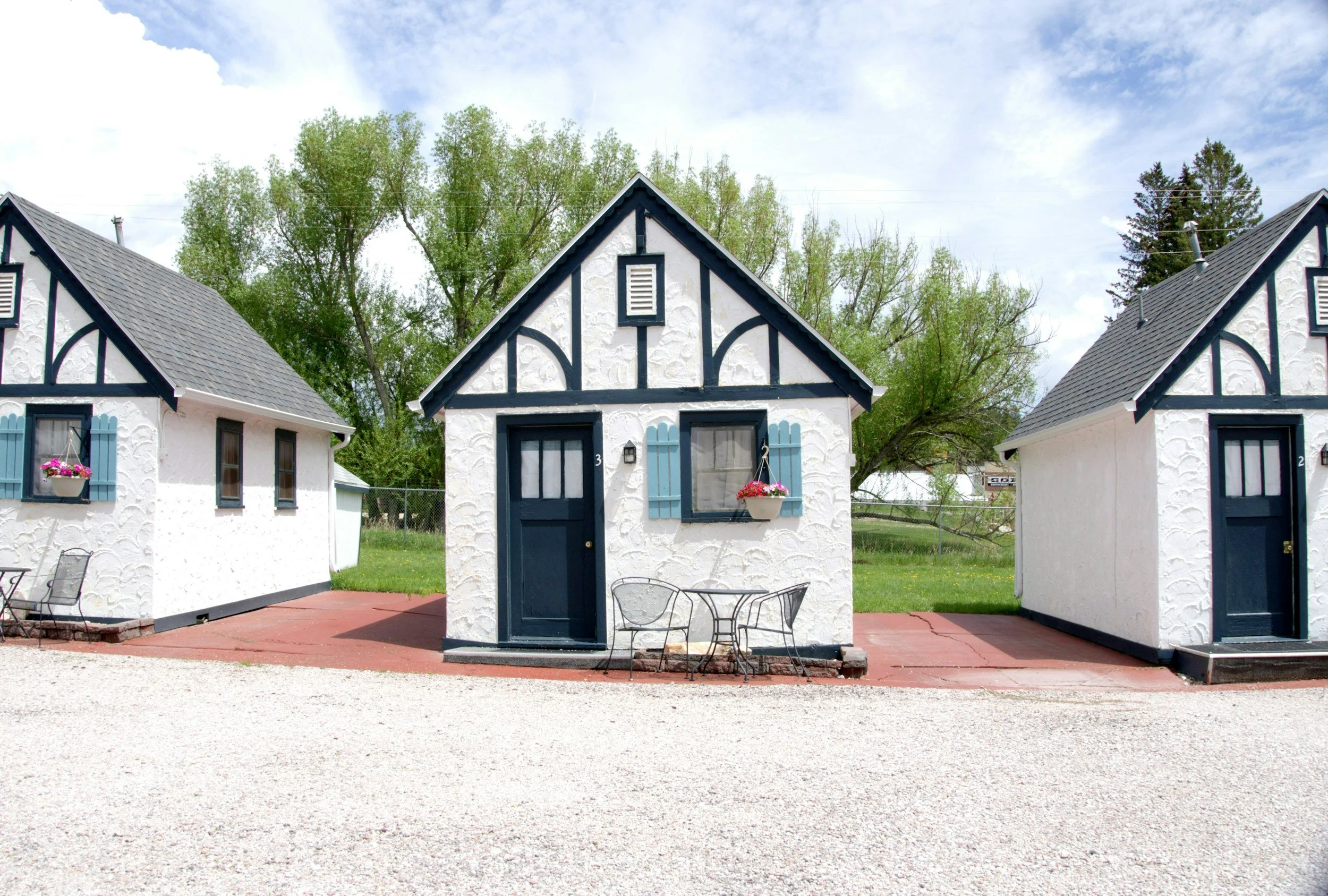 Front view of a white cottage with black trim, blue shutters, and a gabled roof, surrounded by green trees under a partly cloudy sky.