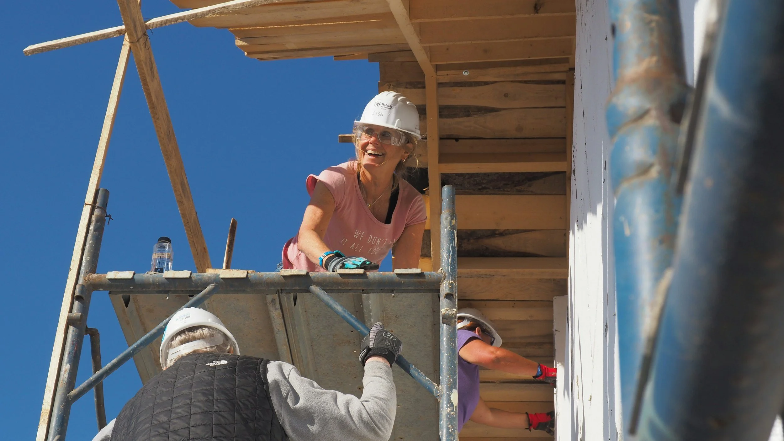 Women working on a construction site, standing on scaffolding, with wooden structure behind and a clear blue sky above.
