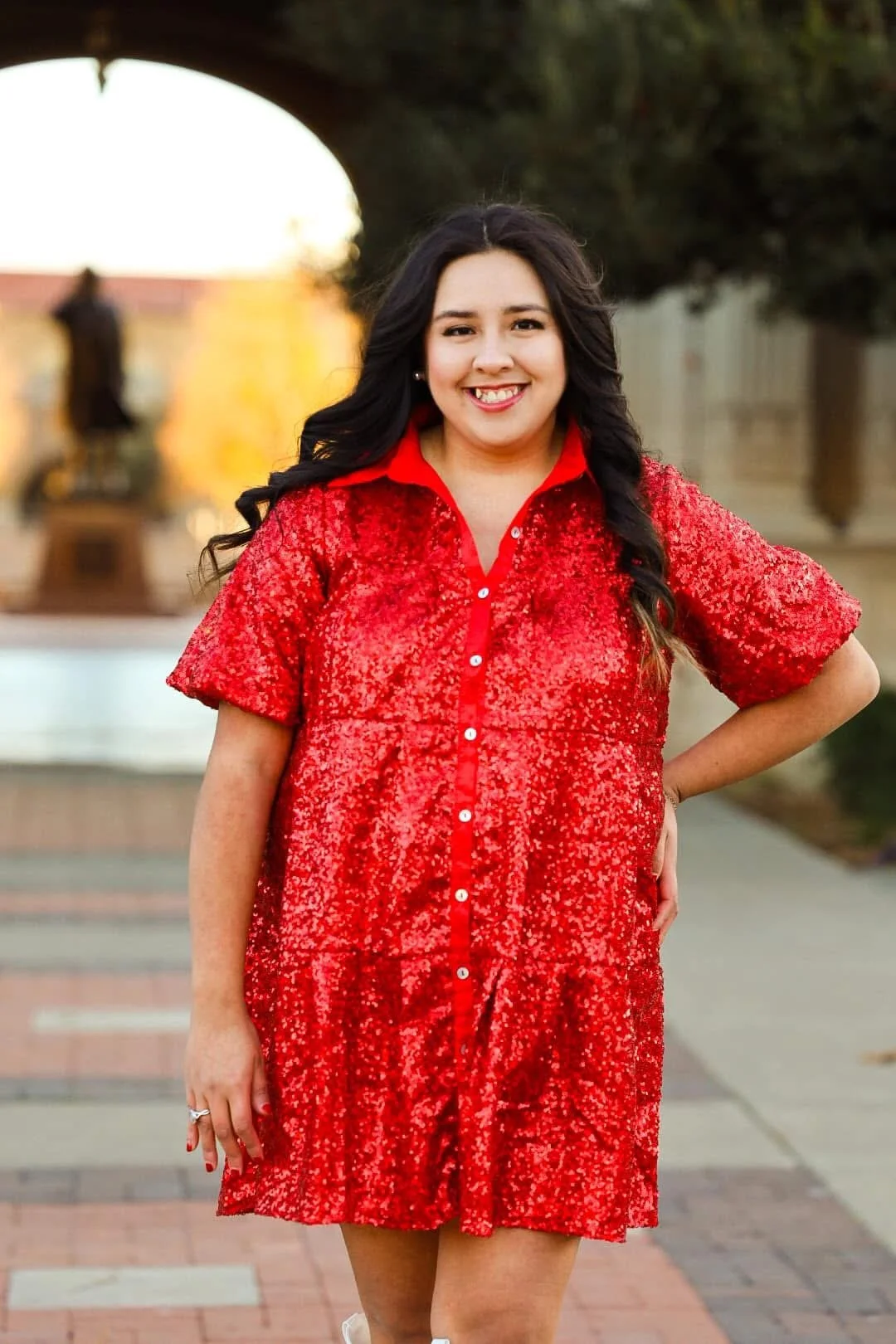Portrait of Molly Ramirez smiling outdoors in a red sequined dress, standing on a brick walkway beneath an archway.