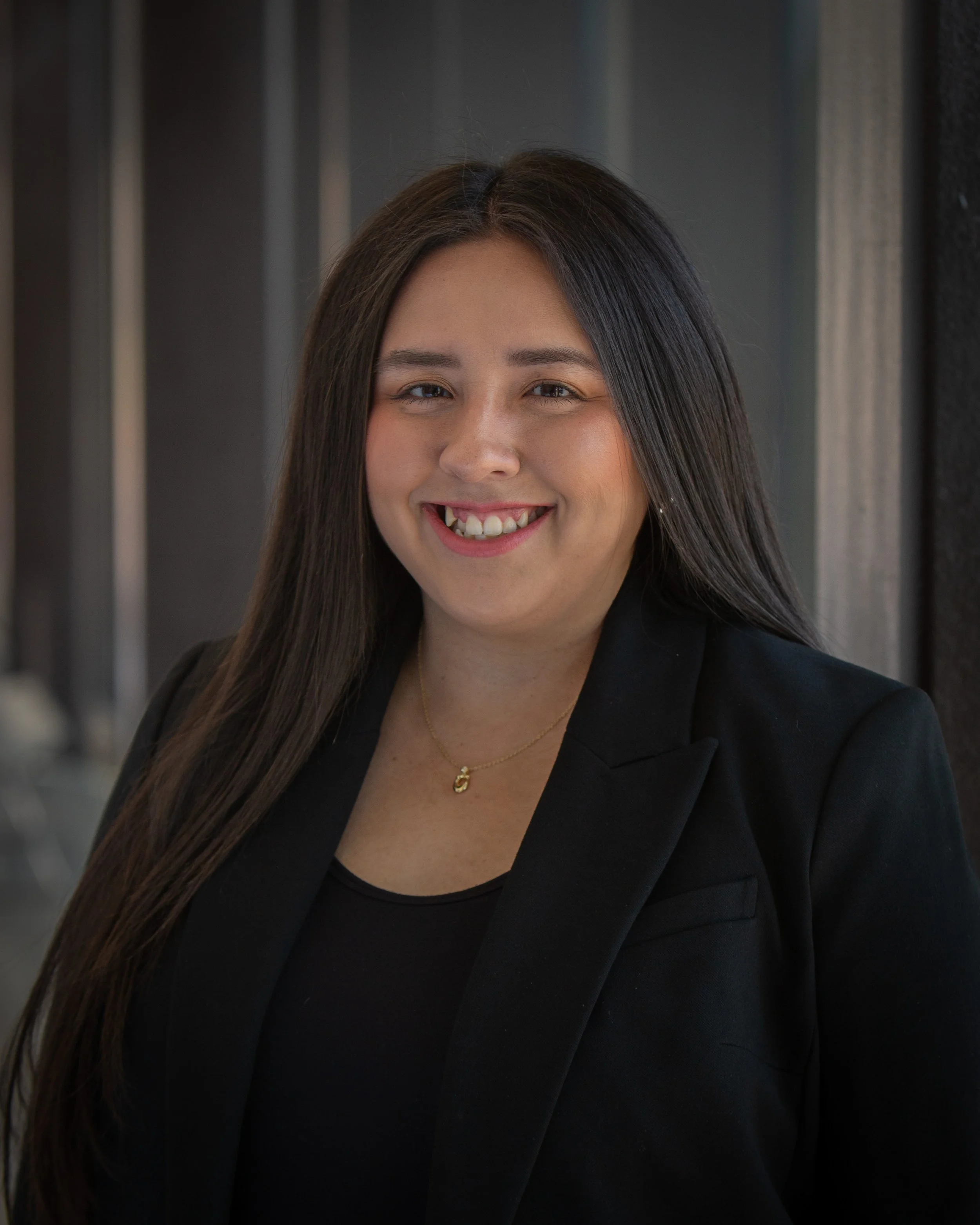 Molly Ramirez smiles at the camera in a professional headshot, wearing a black blazer and black top, with a softly blurred architectural background.