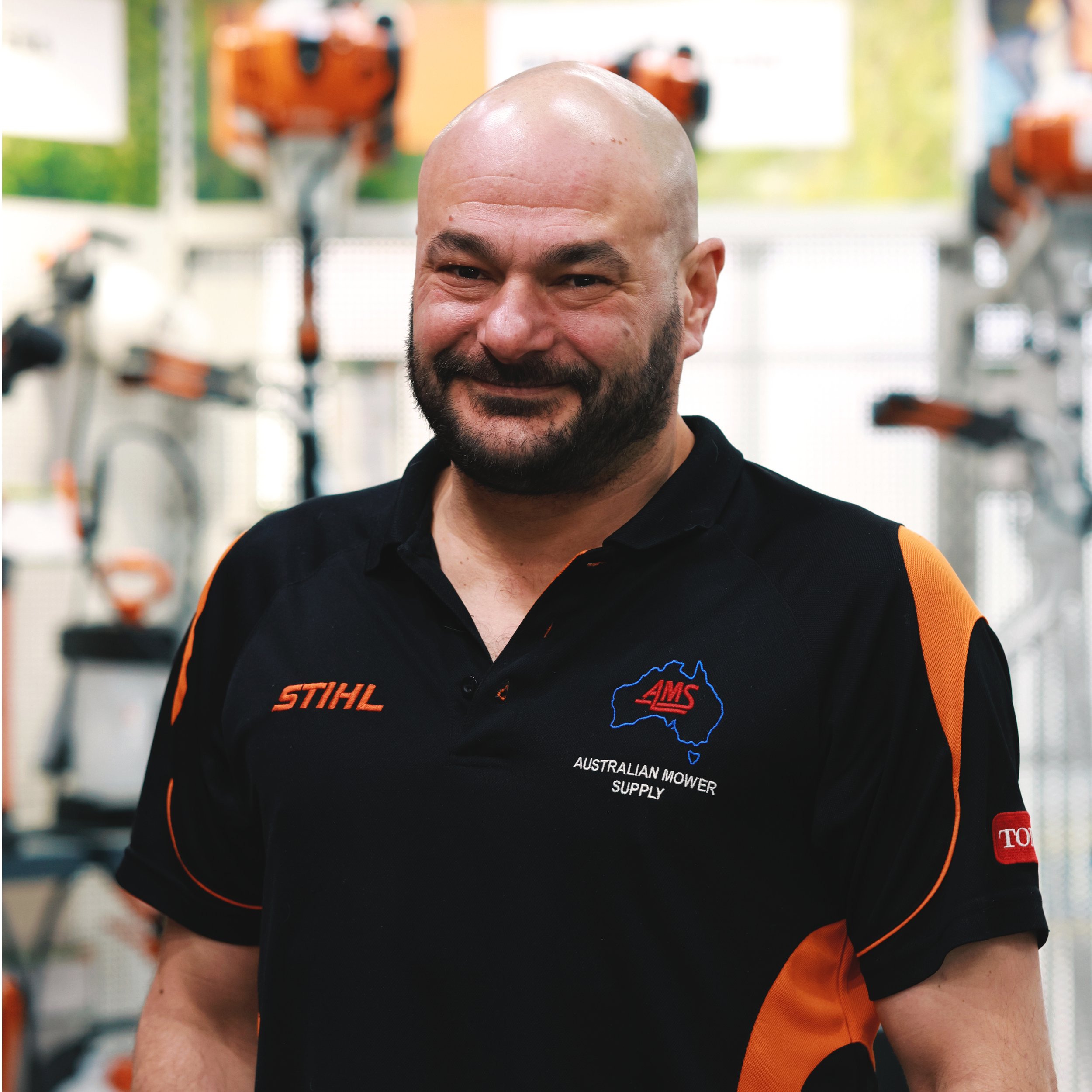 A smiling man with a shaved head and beard, wearing a black sports shirt with orange accents and logos, in a gym or workshop with orange and black power tools in the background.