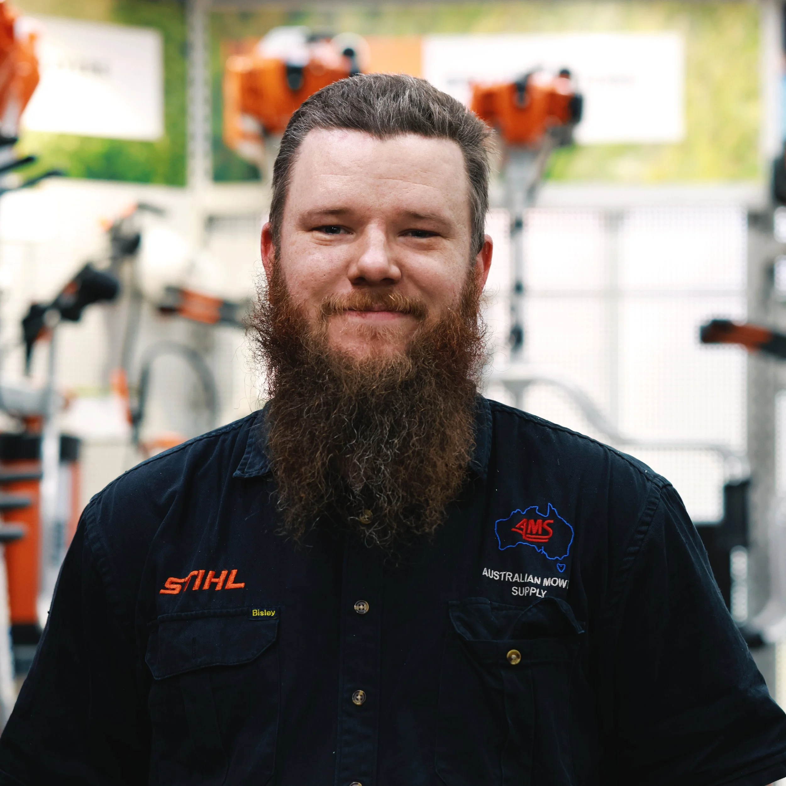 Close-up of a man with a beard, wearing a black work jacket with logos, standing in a workshop with power tools and equipment in the background.