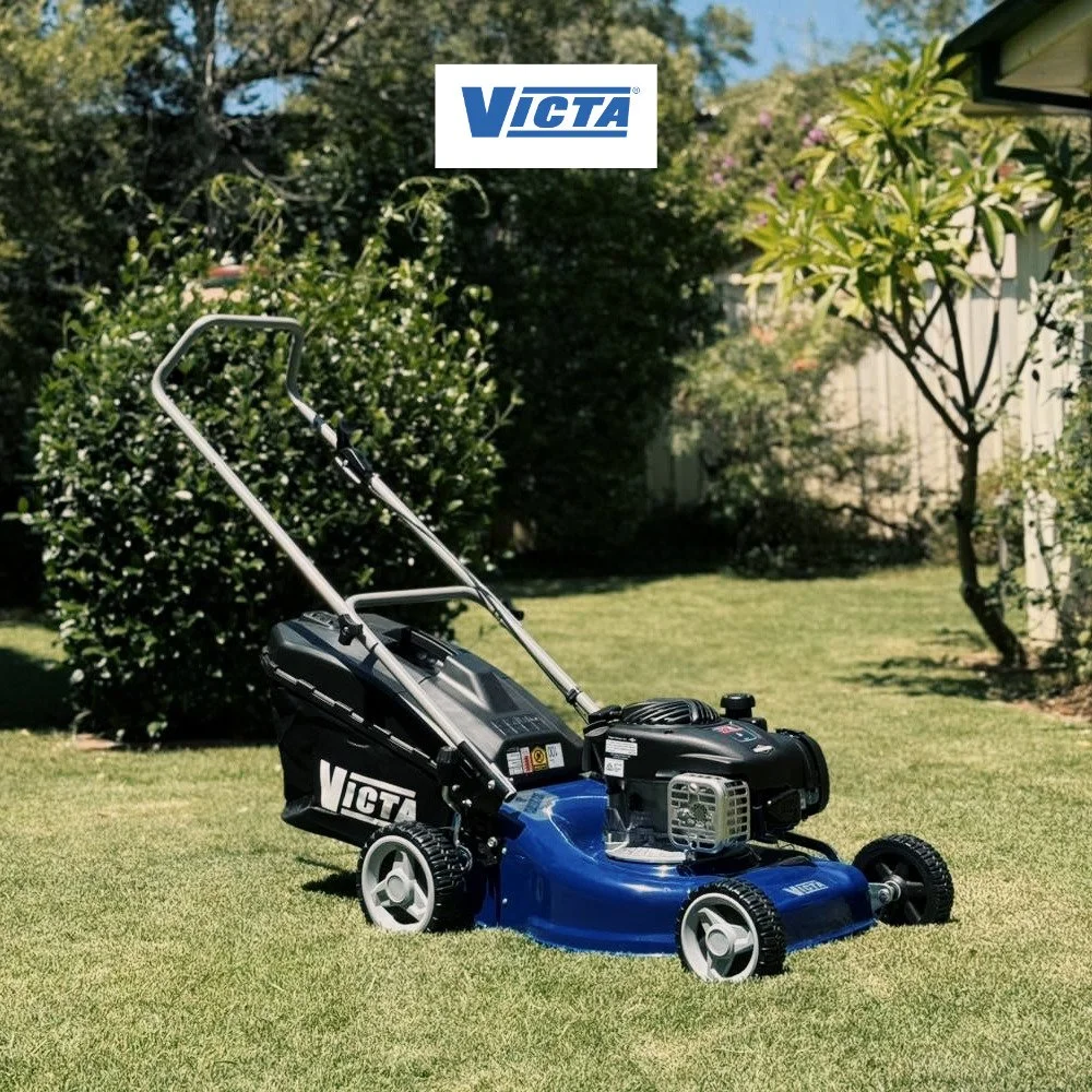 A VICTA brand gasoline-powered lawn mower on a well-maintained grassy lawn with trees and bushes in the background.
