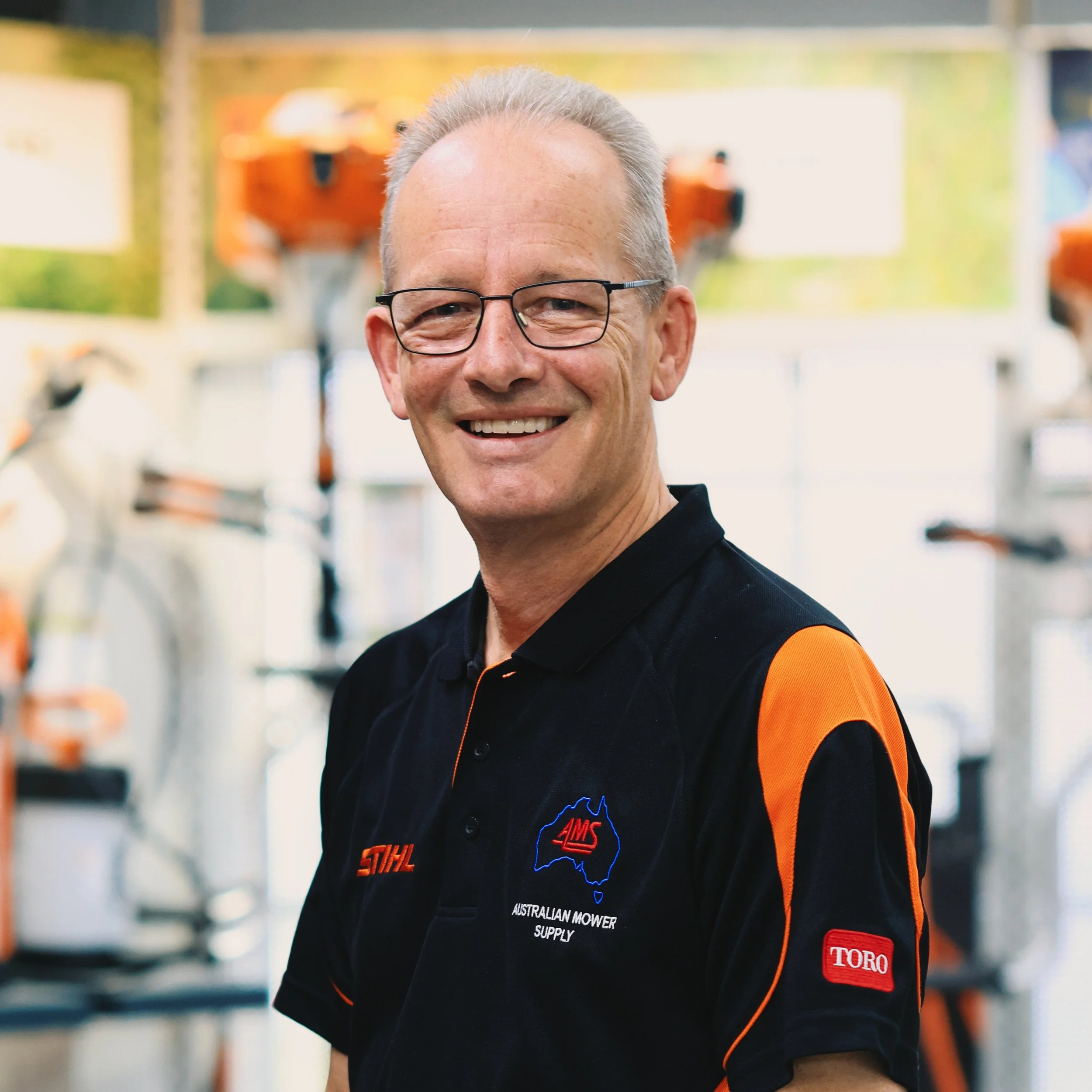 Smiling man wearing glasses and a black uniform with orange accents, with logos of Australia and Toro, standing in a hardware or toolbox aisle.