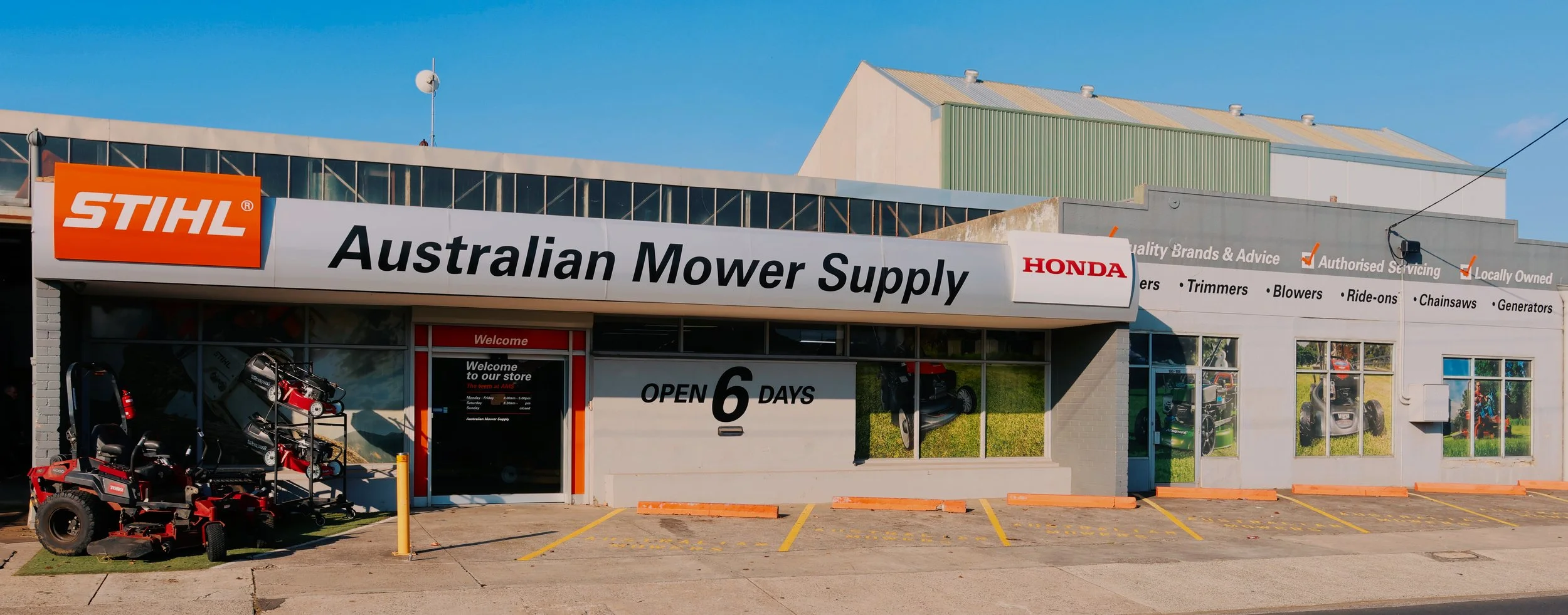 Outside view of a store named Australian Mower Supply, selling outdoor equipment. Store signage includes brands like STIHL and Honda, and lists services such as trimmers, blowers, ride-ons, chainsaws, and generators. The store is open six days a week, with lawn mowers and equipment displayed outside.