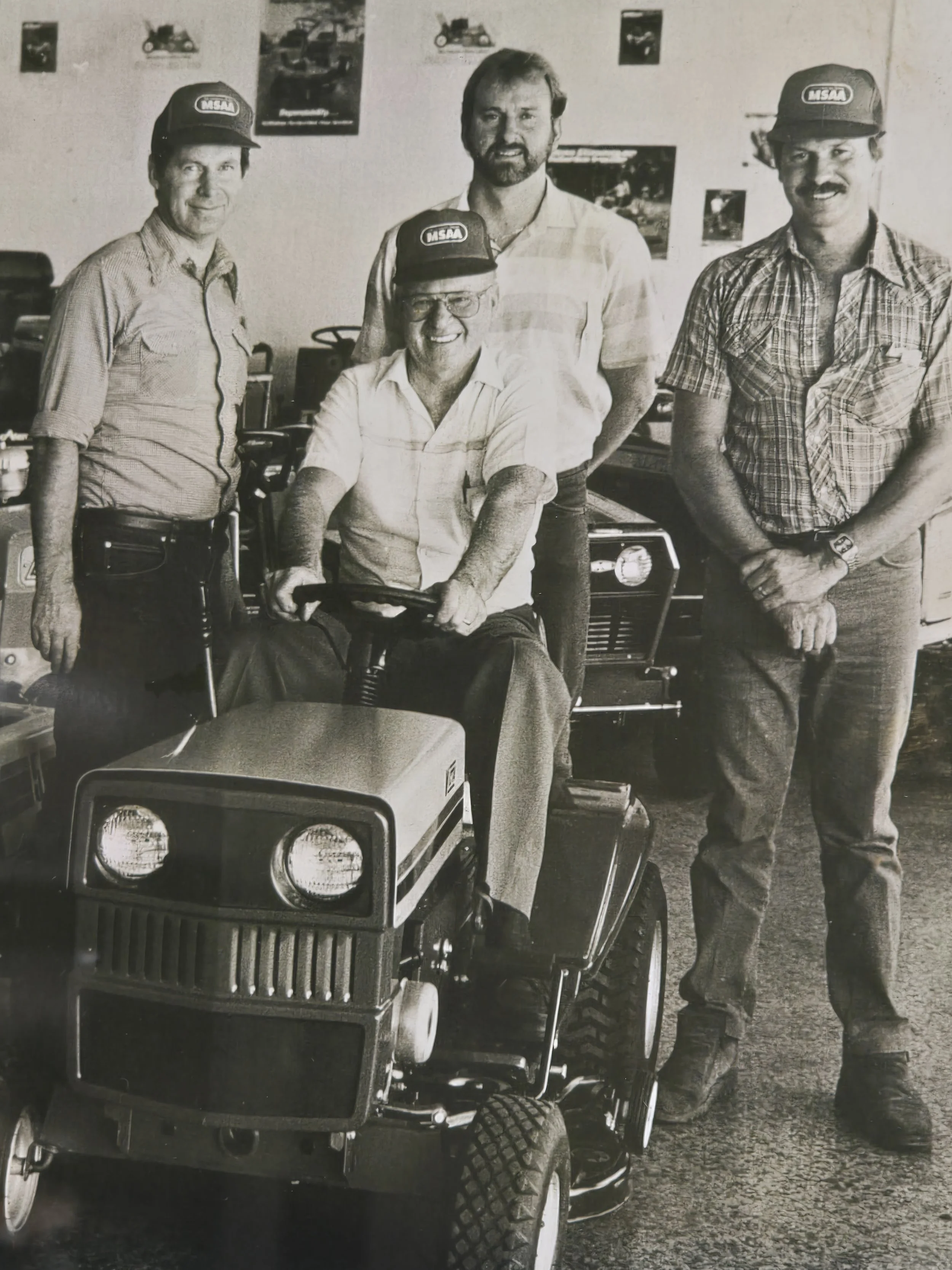 Black and white photo of four men, three standing and one sitting on a small motorized vehicle. All wear hats with the MSAA logo, casual shirts, and jeans. The men are smiling and posing indoors with posters and photos of vehicles on the wall behind them.