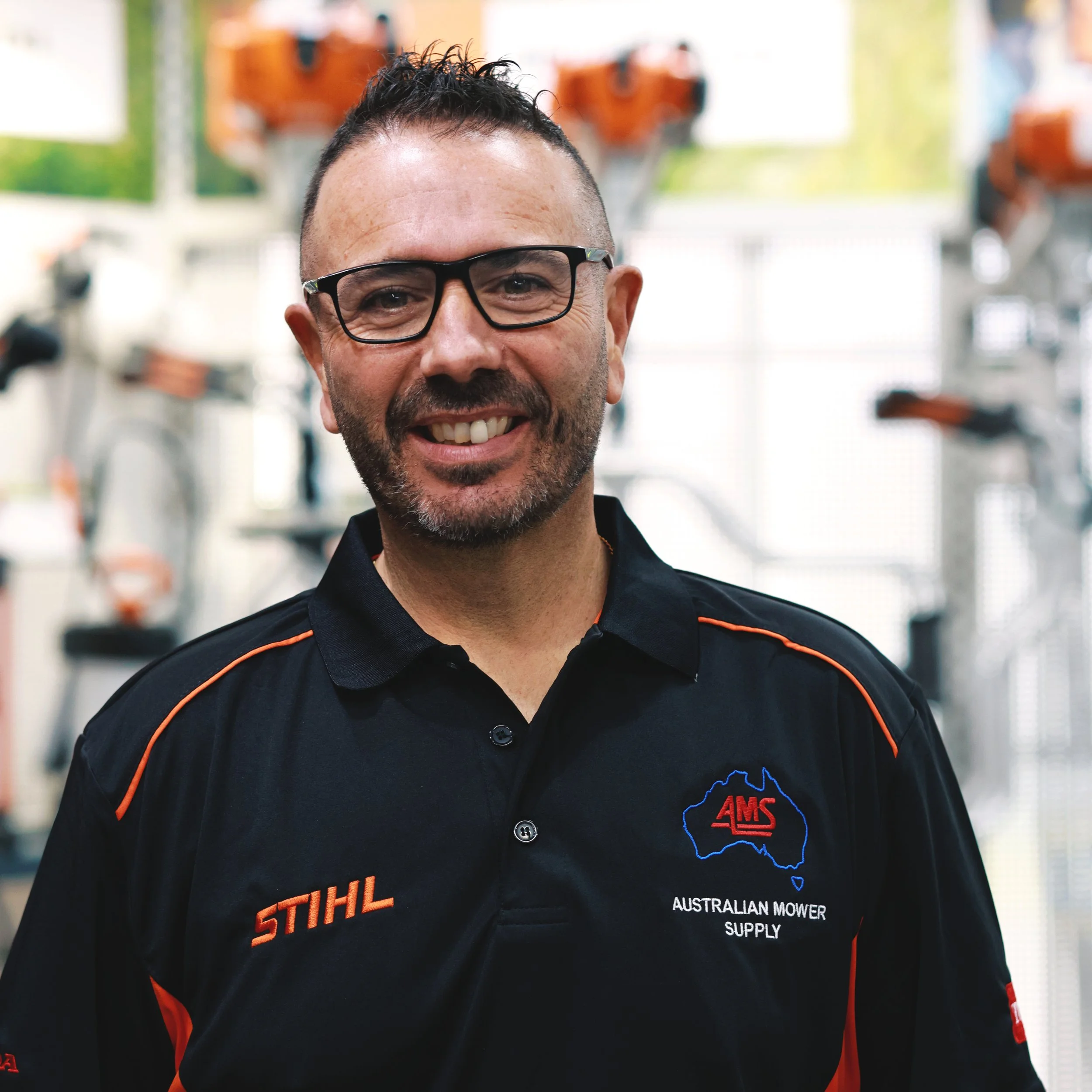Close-up photo of a smiling man with glasses, facial hair, and a short hairstyle, wearing a black shirt with orange accents and logos, in an indoor setting with blurred equipment or machinery in the background.