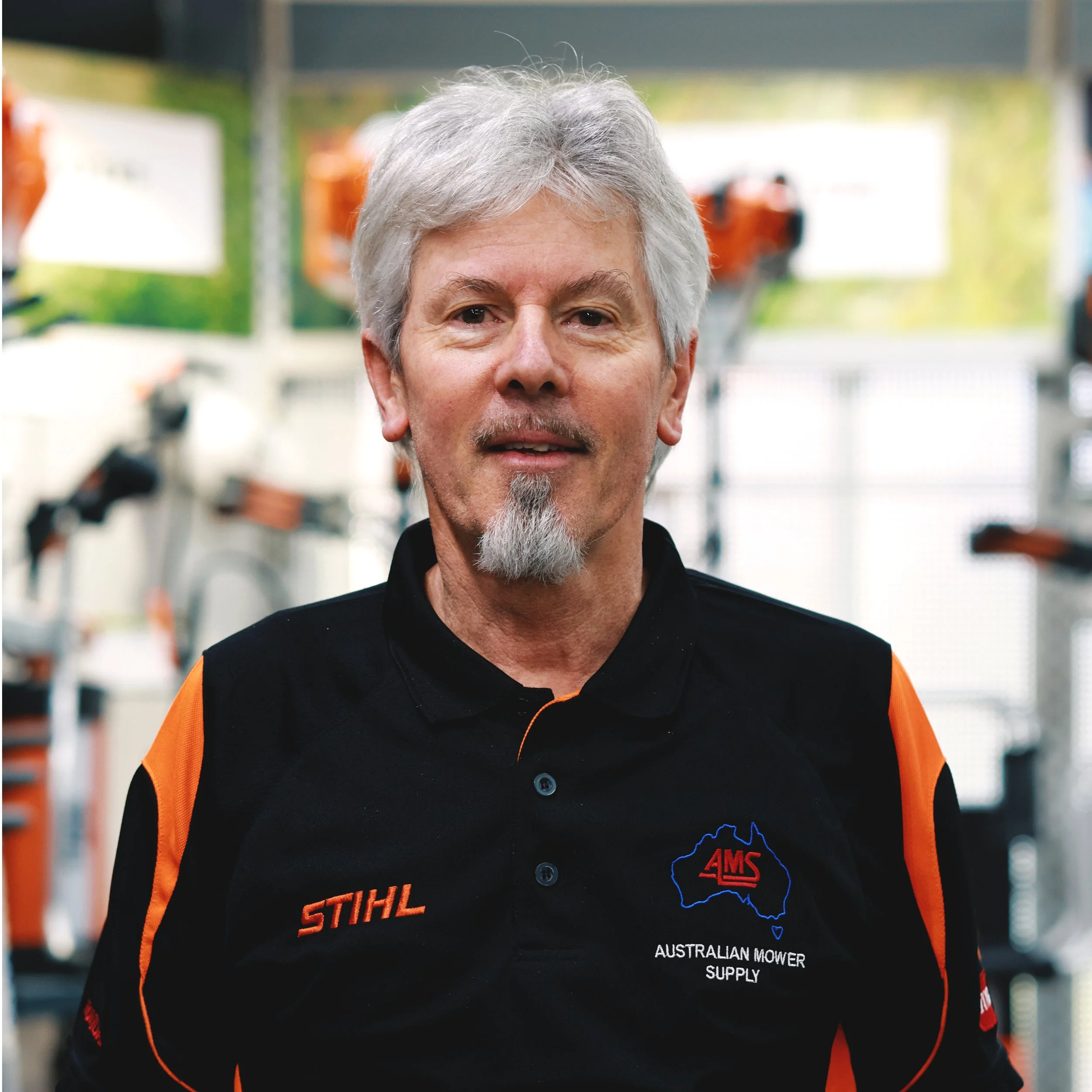 Close-up of a middle-aged man with gray hair and a goatee, wearing a black and orange uniform with logos related to Australian lawnmowers, standing indoors with a blurred background of equipment.