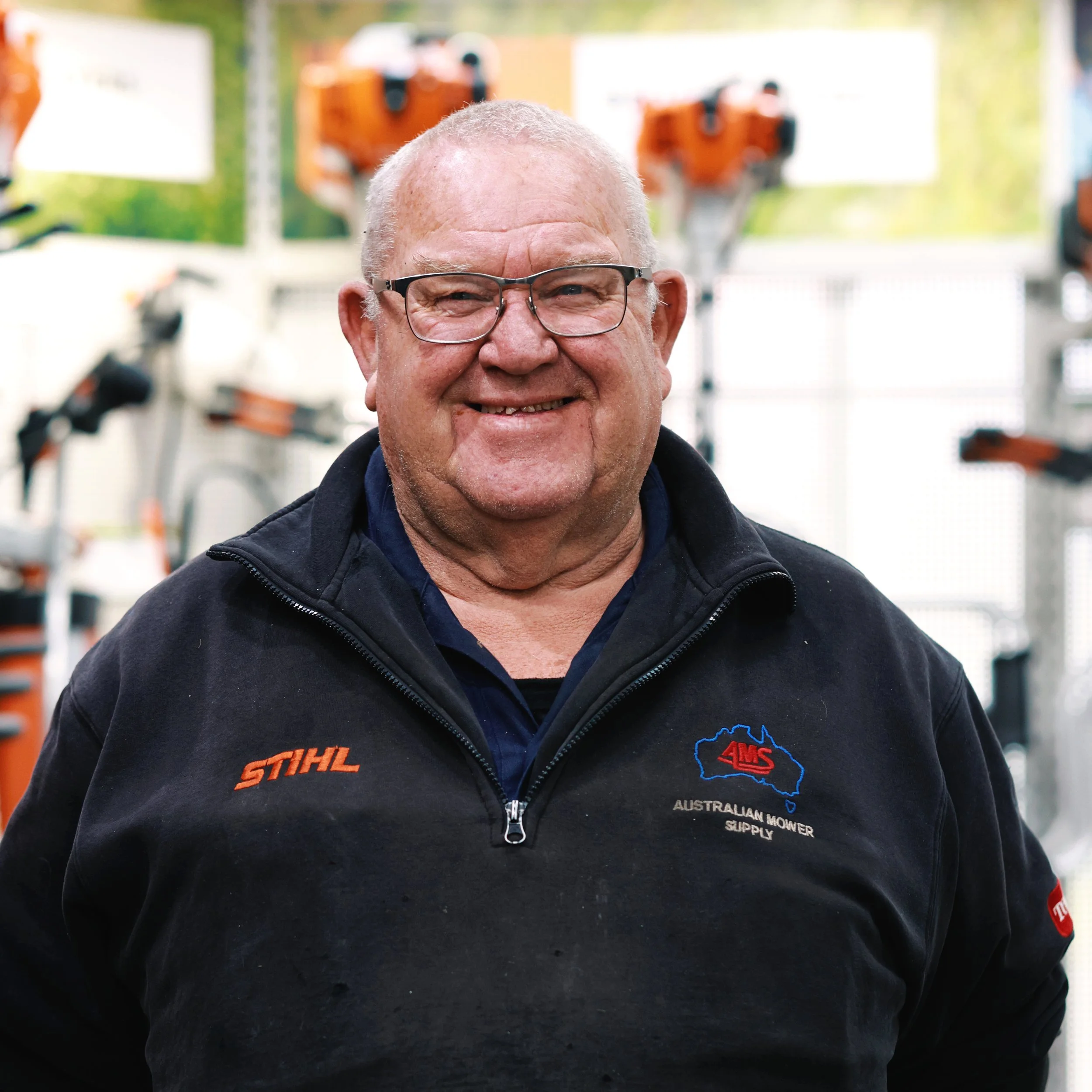 Close-up of an older man with glasses smiling, wearing a black jacket with logos, standing outdoors with a blurred background of machinery.