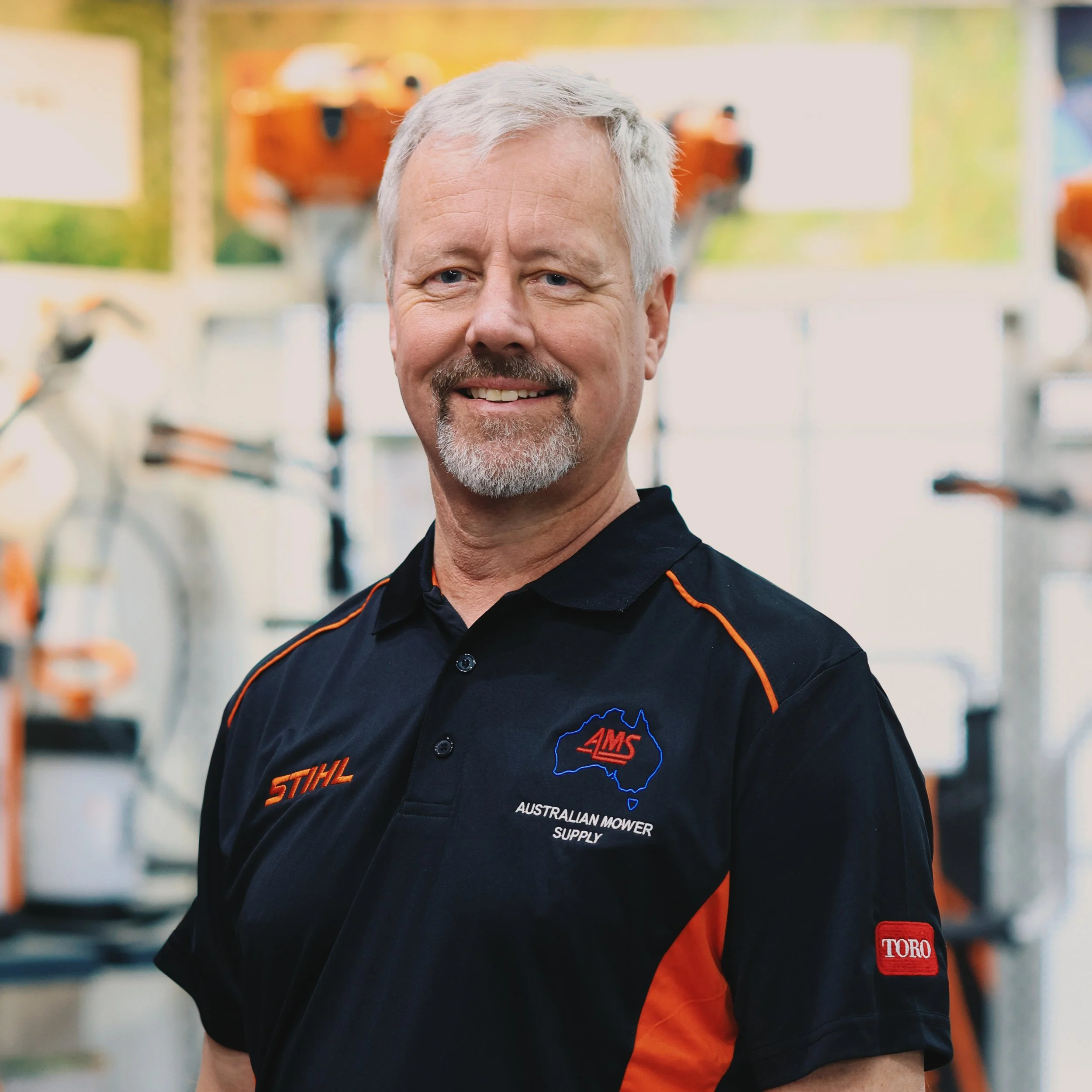 A smiling man with gray hair and a beard wearing a black and orange work shirt with logos, standing outdoors with blurred greenery and equipment in the background.