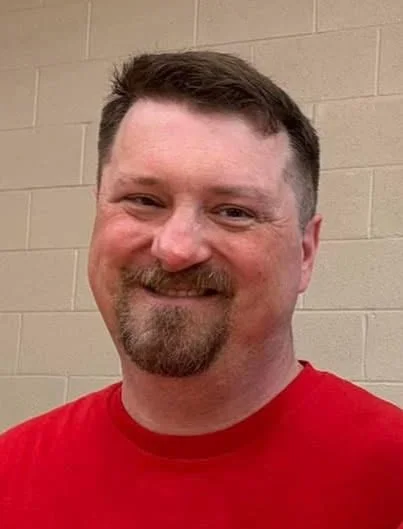A smiling man with short brown hair and a beard, wearing a red shirt, standing in front of a light-colored brick wall.