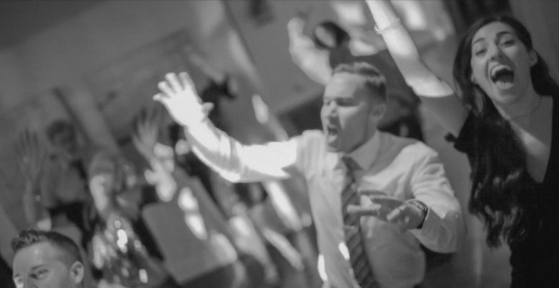 People at an event, laughing and raising their hands, with a woman in black and a man in a suit among the crowd.