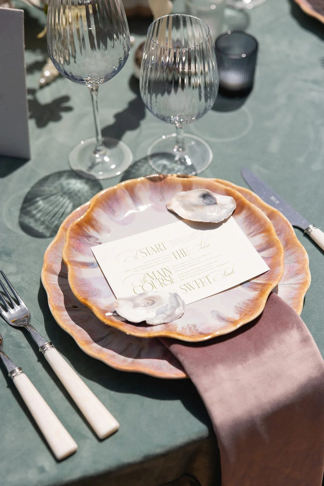 Table setting with a pink scalloped ceramic plate holding an event menu, with two oysters and a pink napkin underneath. Silverware with white handles, two empty crystal wine glasses, and a black candle holder in the background. wedding tabletop items
