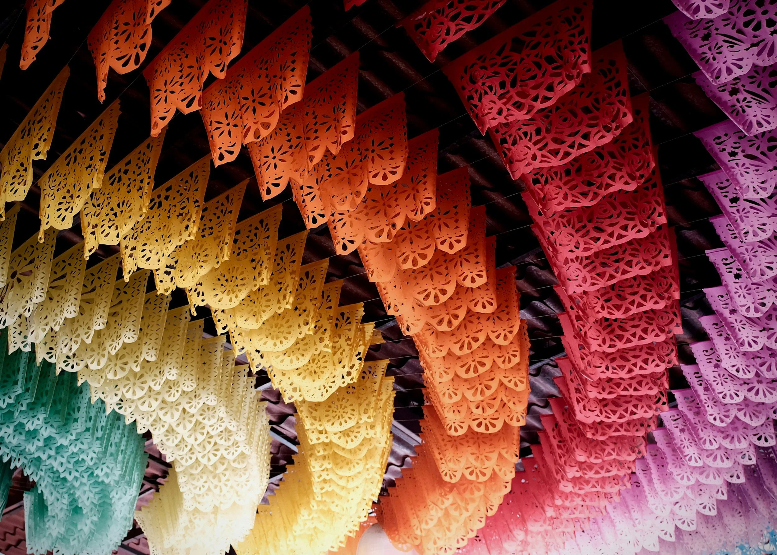 Colorful papel picado banners hanging, in yellow, orange, red, and purple, with intricate cut-out designs, typically used for festive decorations.