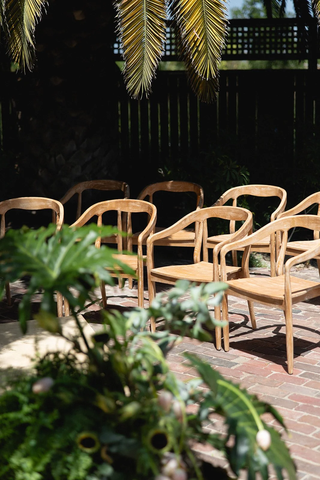 Multiple wooden chairs arranged outdoors on a brick patio, surrounded by greenery and shaded by palm tree leaves. wedding ceremony. luxury. high end.