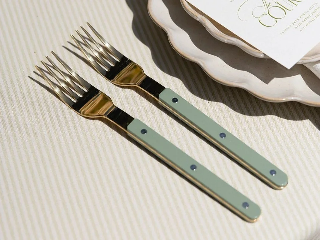 Two forks with light green handles on a striped tablecloth, near a decorative plate with a menu or invitation card on top. luxury tabletop items. luxury wedding