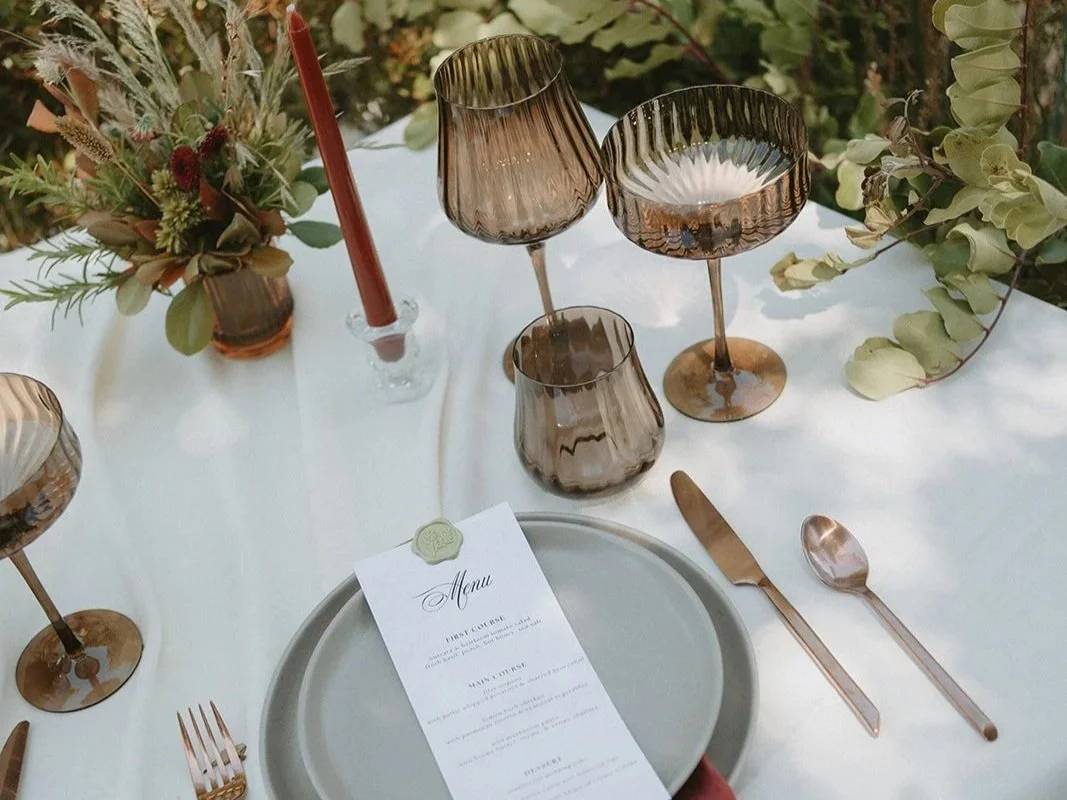 Elegant dining table setup with amber glassware, a white menu, and a floral centerpiece arranged with greenery and dried flowers.
