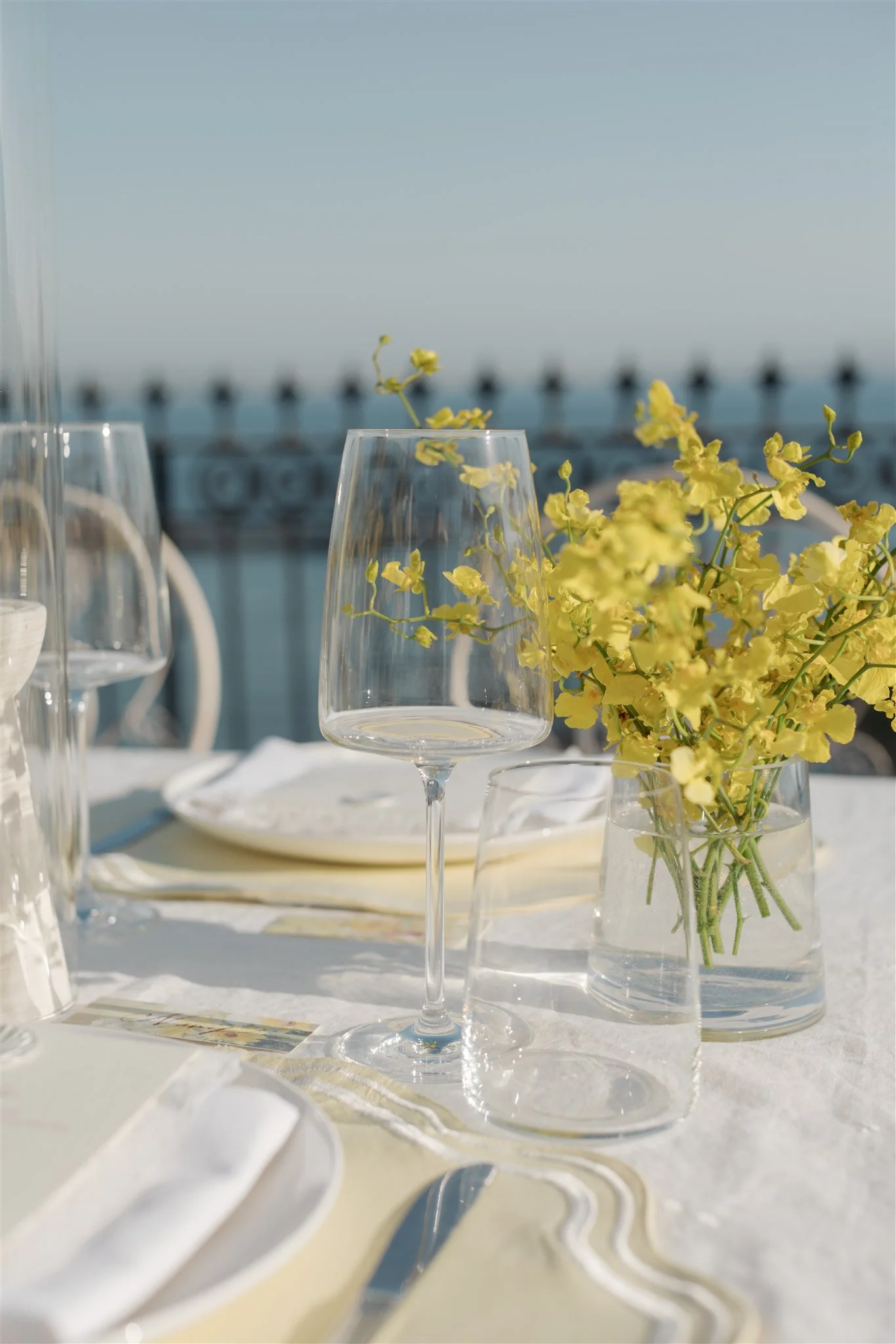 A table set for an outdoor meal with wine glasses, a bunch of yellow flowers in a glass vase, and a napkin. In the background, a railing and a clear sky are visible. wedding tabletop setup