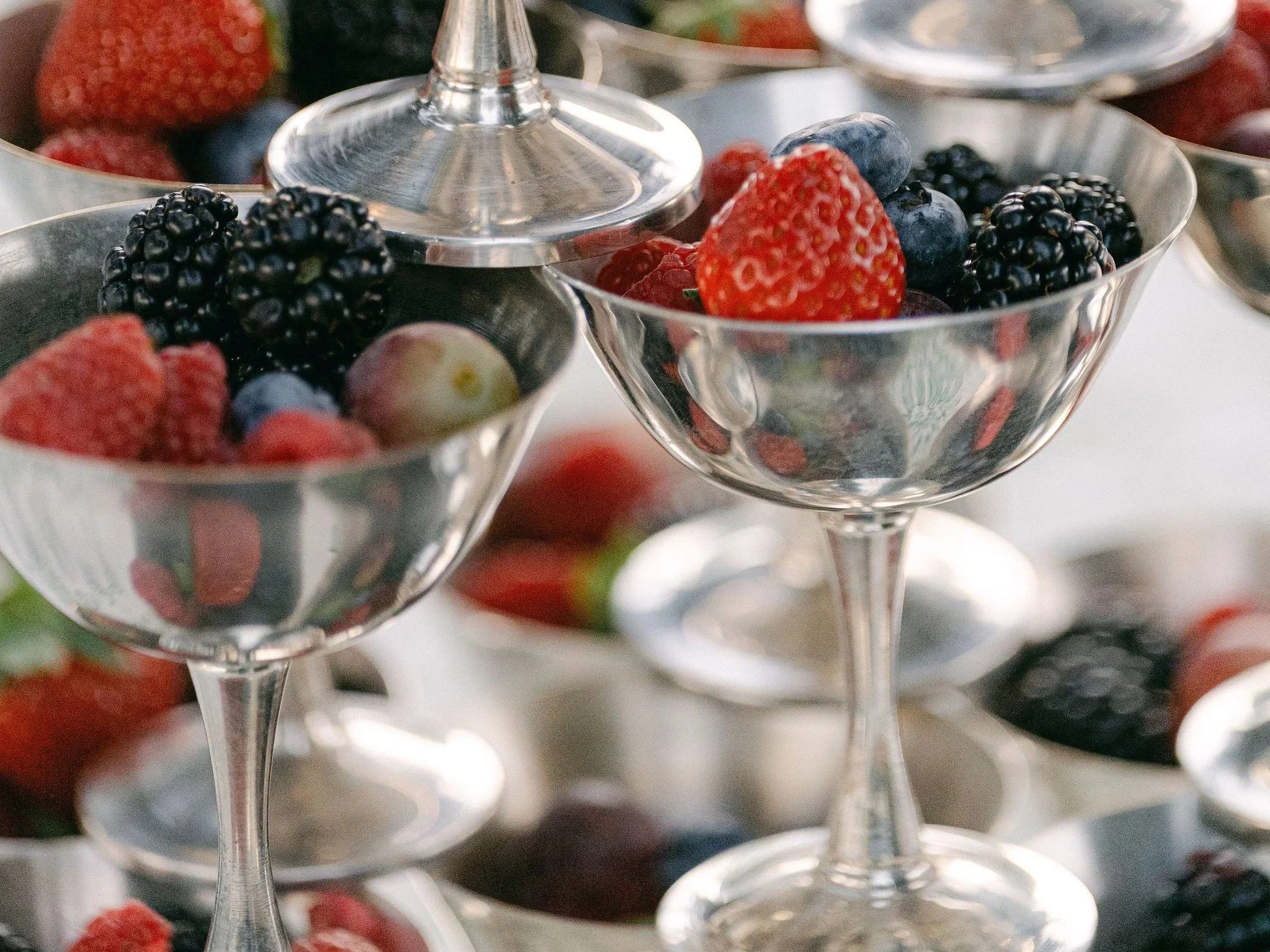 Close-up of glass bowls filled with various fresh berries, including strawberries, blueberries, blackberries, and raspberries, arranged on a tiered serving stand. Coupe cups