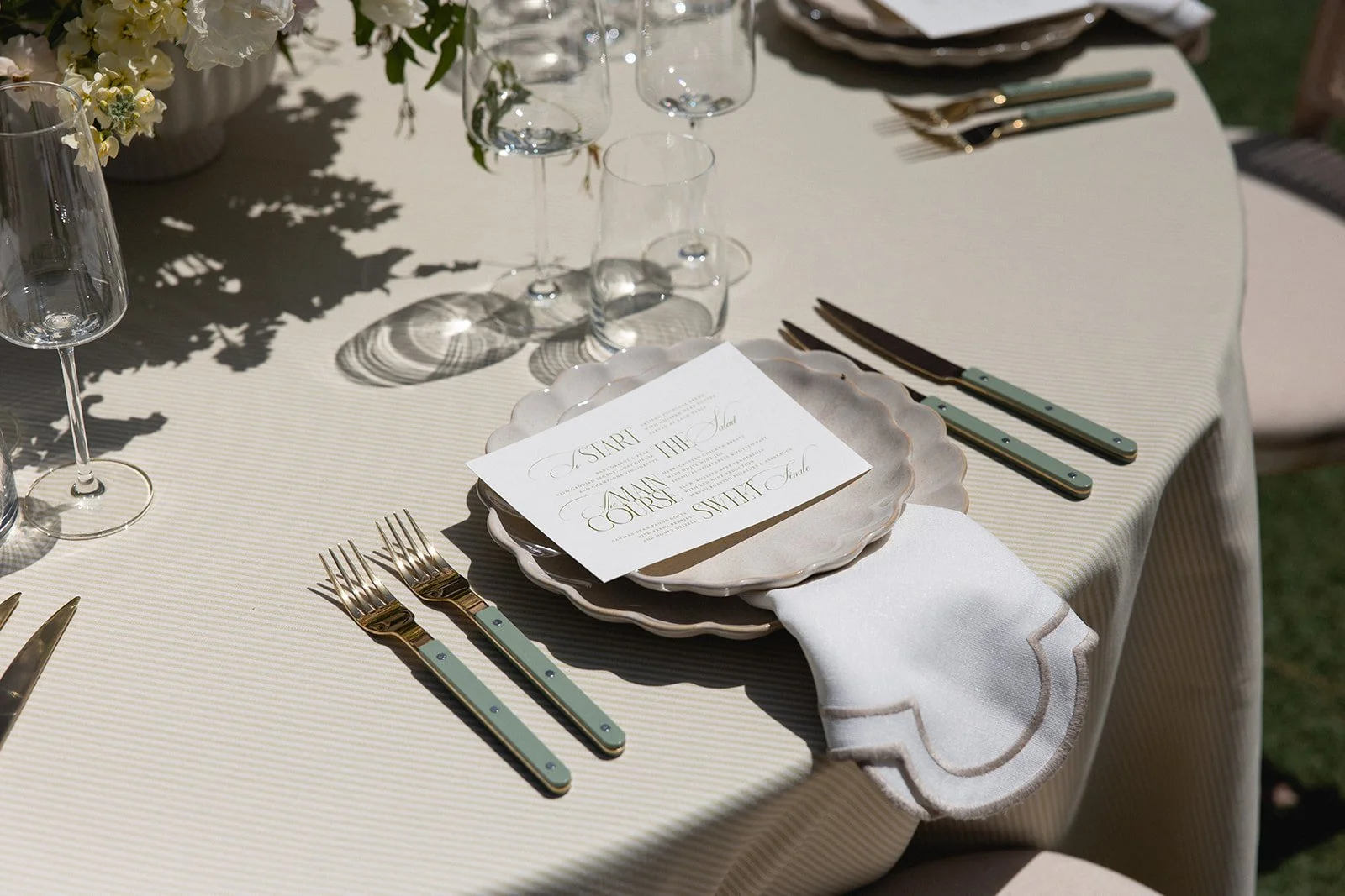 A formal outdoor table setting with plates, silverware, wine glasses, and a printed menu. Plates are stacked, and a white napkin is placed underneath. A floral centerpiece and sunlight casting shadows are visible, indicating a daytime event. wedding