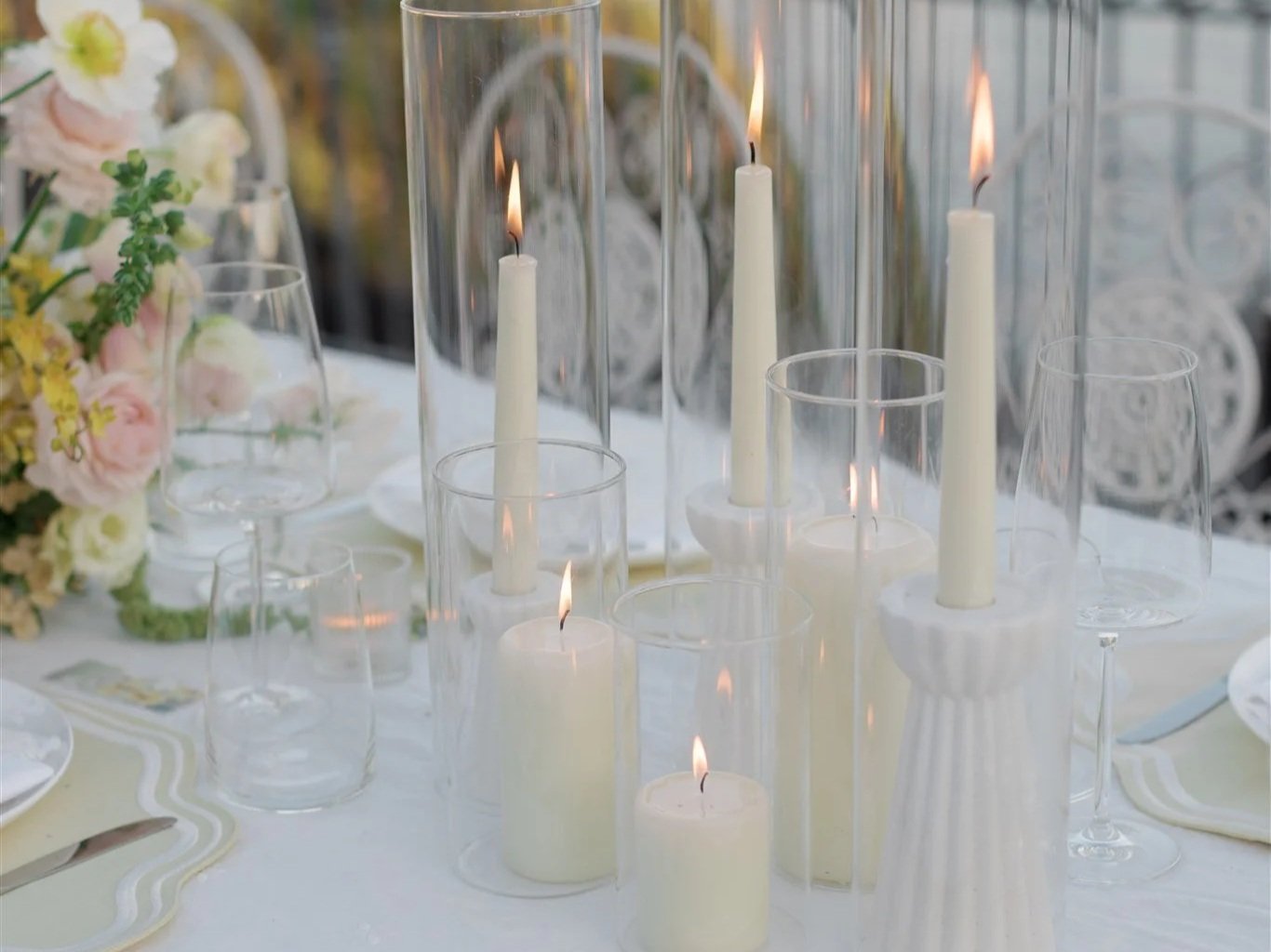 Elegant table setting with multiple lit white candles in glass holders and some empty wine glasses, next to a floral centerpiece with pastel-colored flowers.