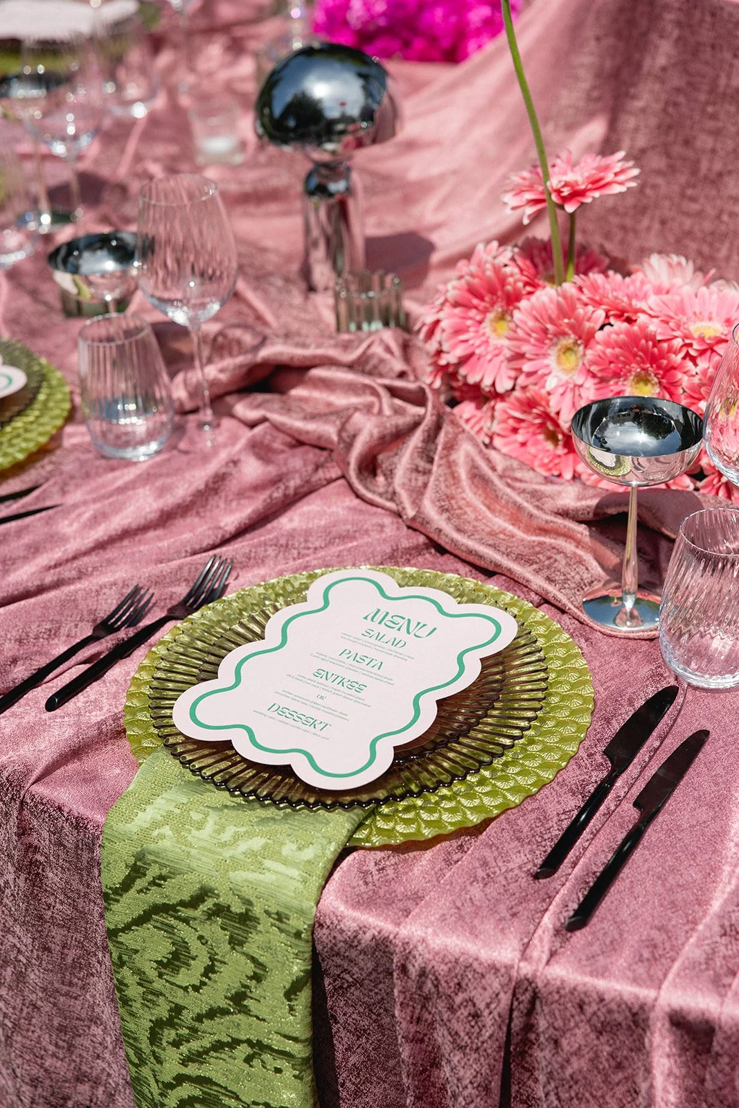 A pink party table decorated with pink flowers, a green textured charger plate, a pink menu card, black cutlery, and glassware. eclectic wedding setup. reception. one of a kind