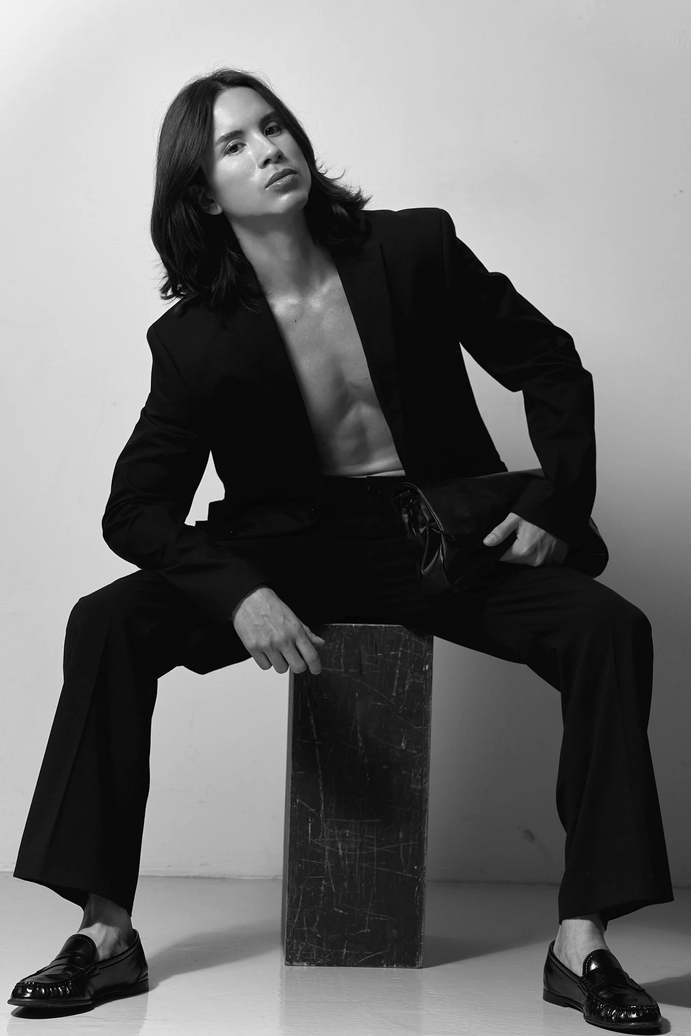 A black-and-white portrait of a woman with shoulder-length dark hair, wearing a blazer, sitting on a chest with legs apart, holding a bag, and wearing loafers.