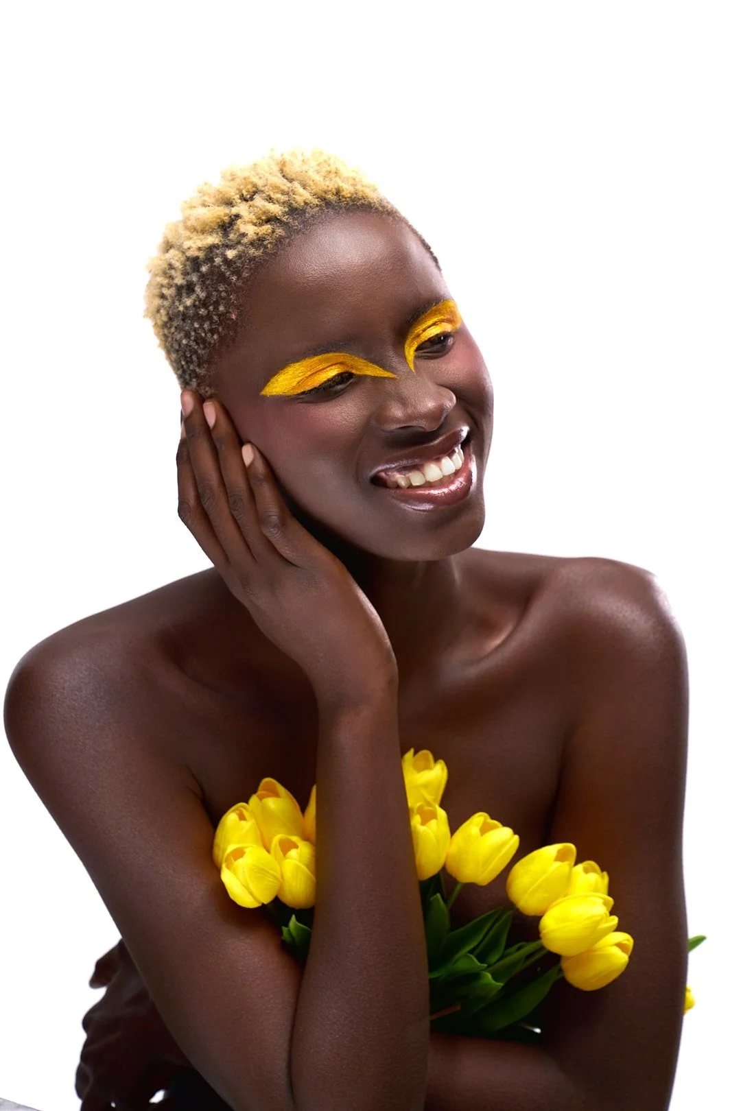 A smiling woman with short blonde hair, yellow eyeshadow, holding a bouquet of yellow tulips, posing against a white background.