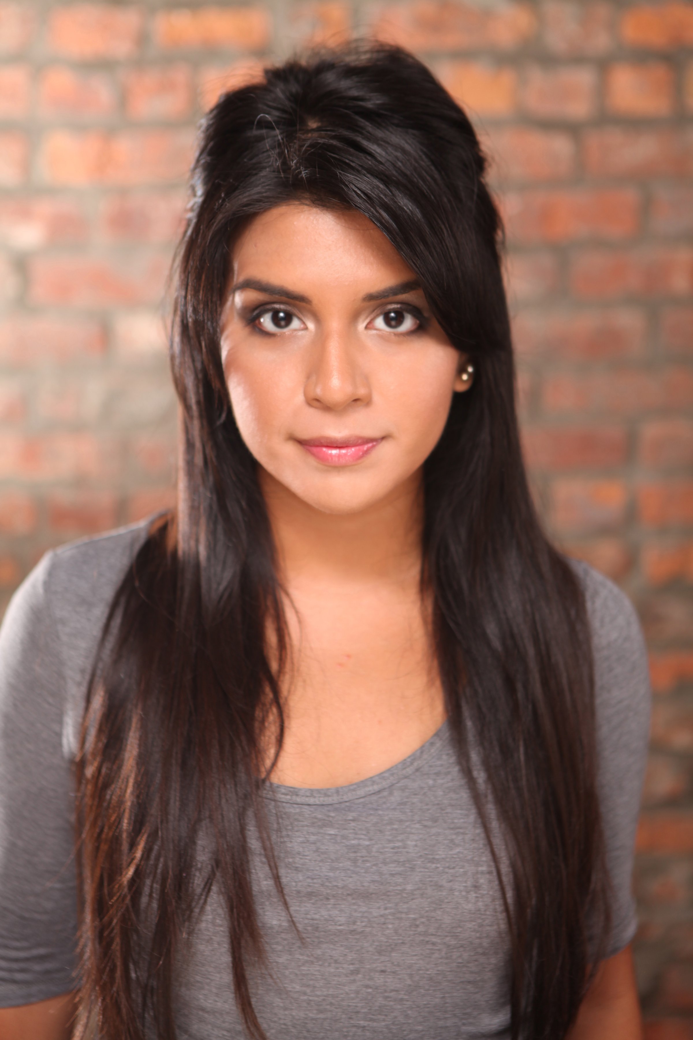 Close-up of a woman with long dark hair and earrings, wearing a gray shirt, standing in front of a brick wall.