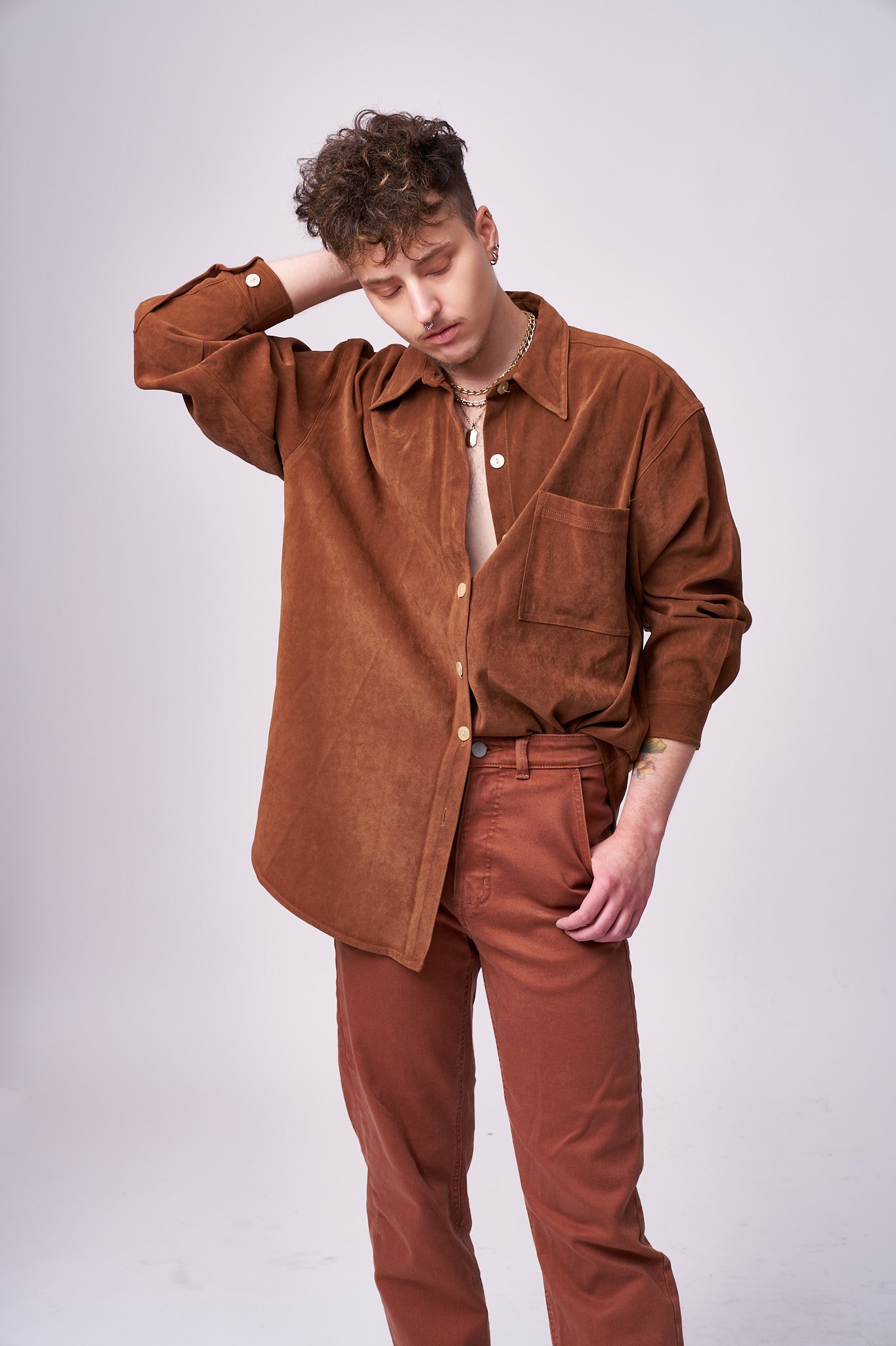 A young man with curly hair and multiple piercings poses with his eyes closed, wearing a brown button-up shirt and matching pants, accessorized with layered necklaces and earrings, standing against a plain light grey background.