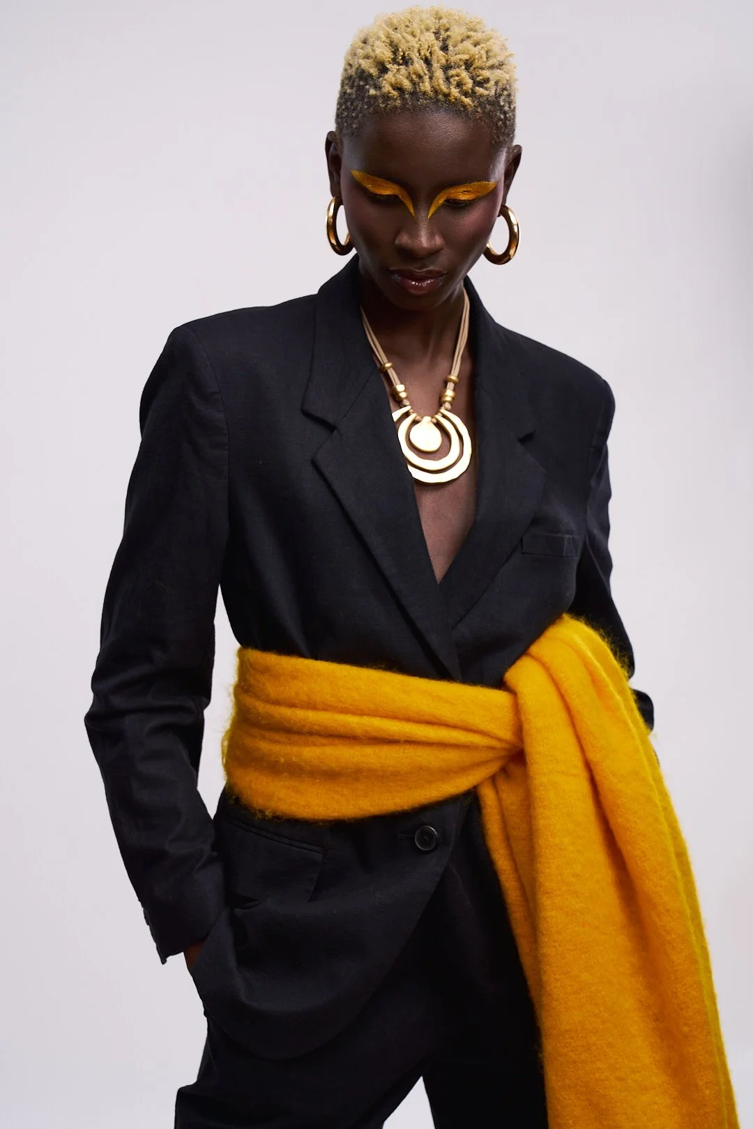 A woman with short, blonde, curly hair styled in a fade. She is wearing a black blazer, large gold hoop earrings, a bold gold necklace, and bright yellow makeup around her eyes. She has a bright yellow scarf tied around her waist and is standing agai