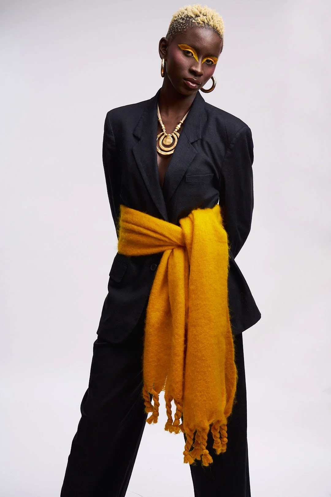 A woman with short blonde hair and dark skin, wearing yellow eye makeup and large hoop earrings. She has on a black blazer with a yellow scarf tied around her waist and a gold necklace, standing against a plain gray background.