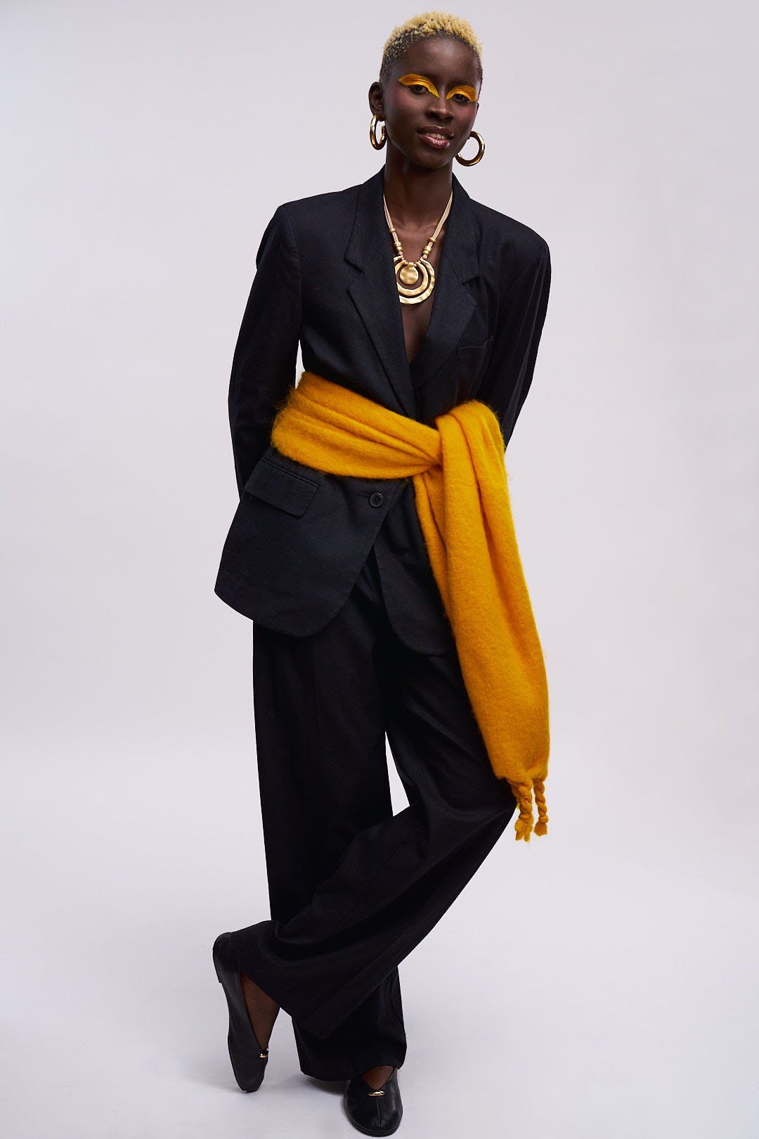 A woman with short blonde hair and dark skin wearing a black suit with a yellow scarf tied around her waist, bold yellow eye makeup, large gold hoop earrings, a gold necklace, and black shoes, standing against a plain white background.