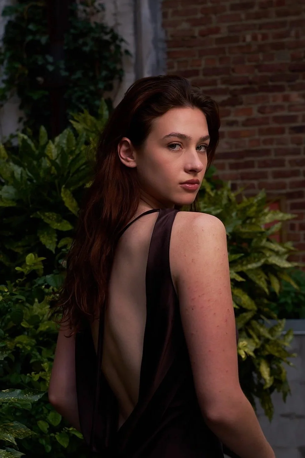 A young woman with long, dark hair looking over her shoulder, standing outdoors near green foliage and a brick wall.