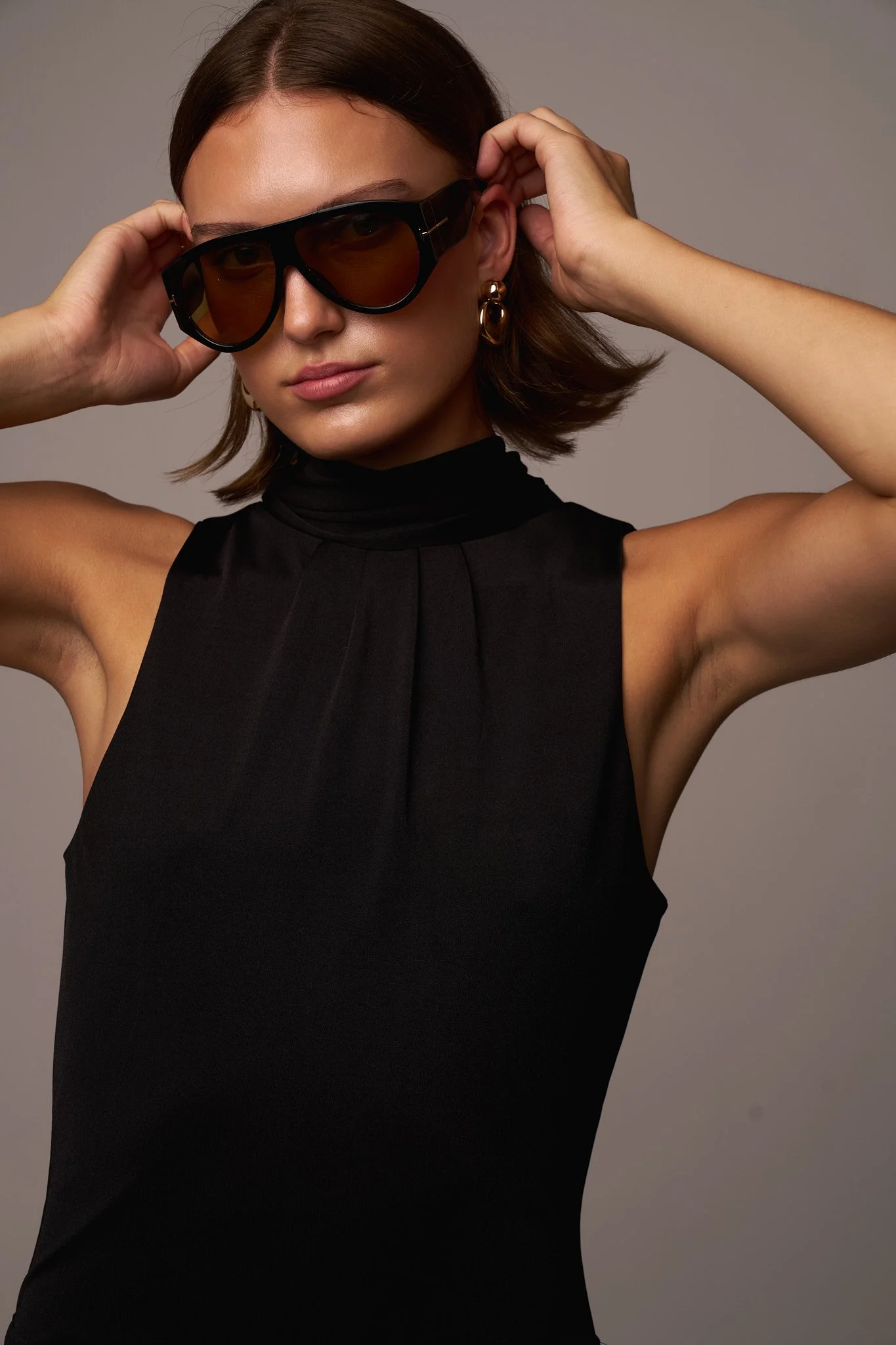 A woman with dark brown hair in a bob hairstyle, wearing large black sunglasses, gold earrings, and a sleeveless black top with a high collar, adjusting her sunglasses with both hands.