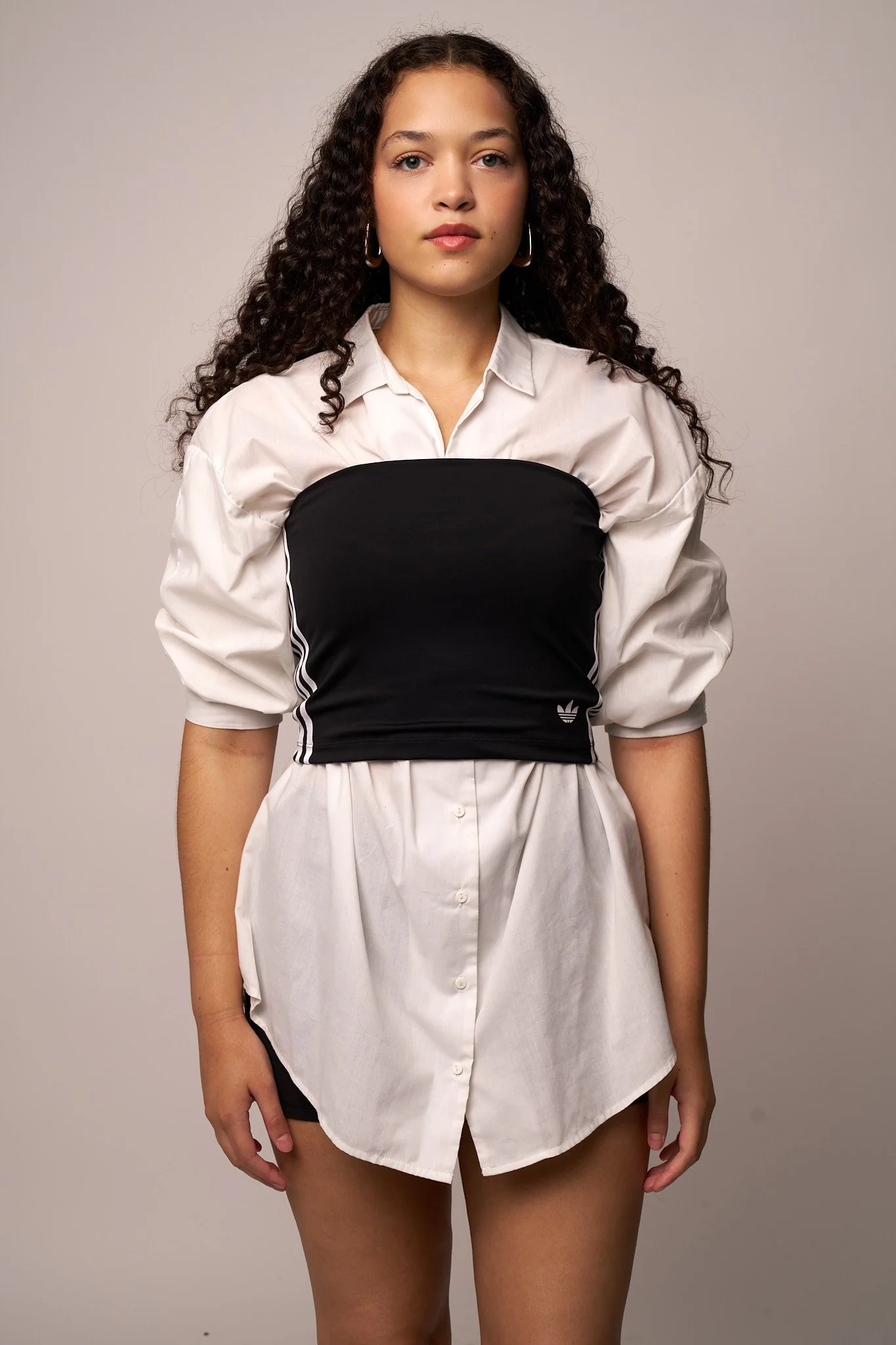 A woman with curly hair wearing a white button-up shirt over a black Adidas sports bra, with a white skirt and black shorts, standing against a plain gray background.