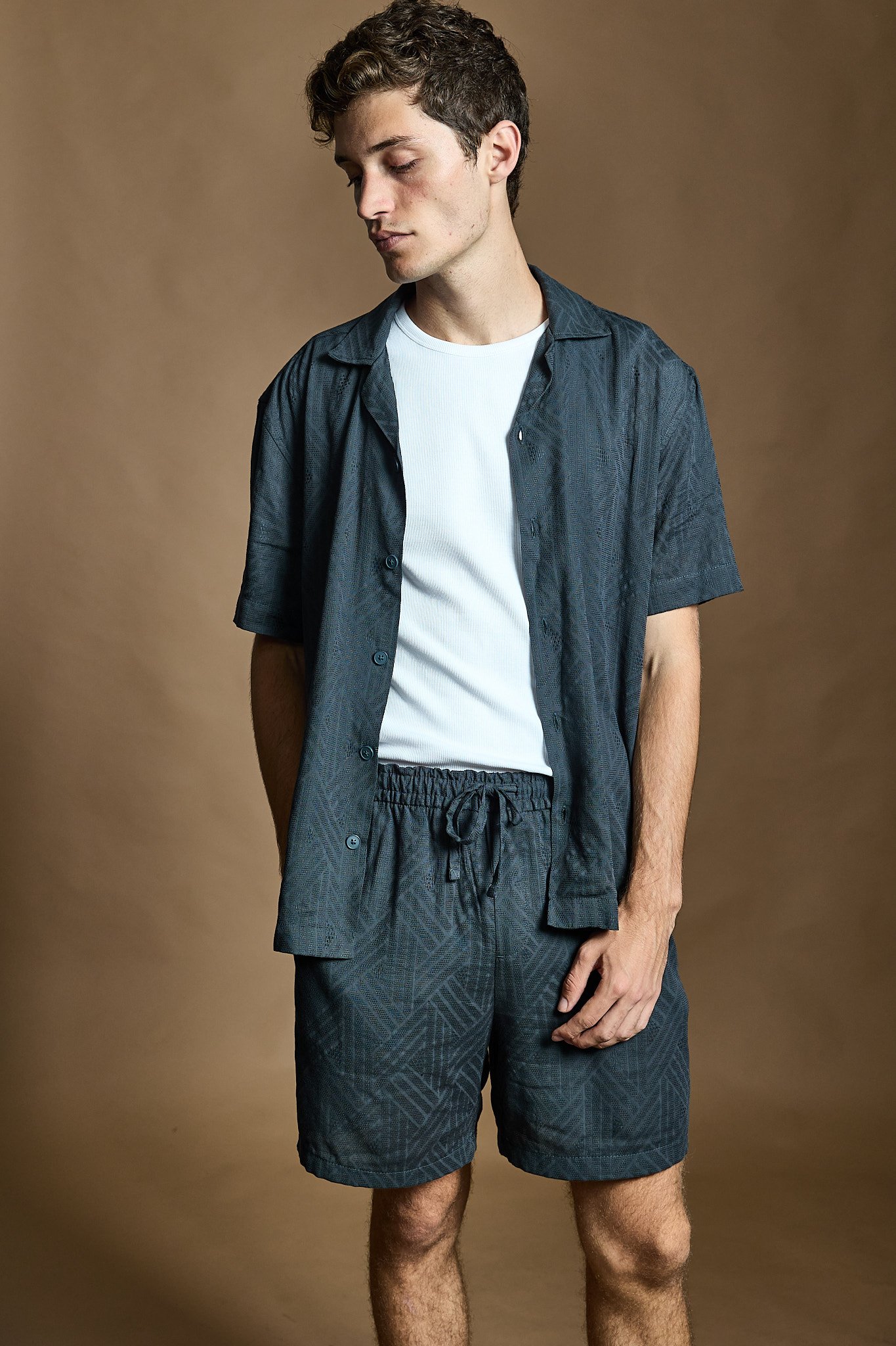 A young man with curly brown hair wearing a white t-shirt, a dark short-sleeve button-up shirt, and matching shorts, standing against a plain brown background with his eyes closed and one hand at his side.