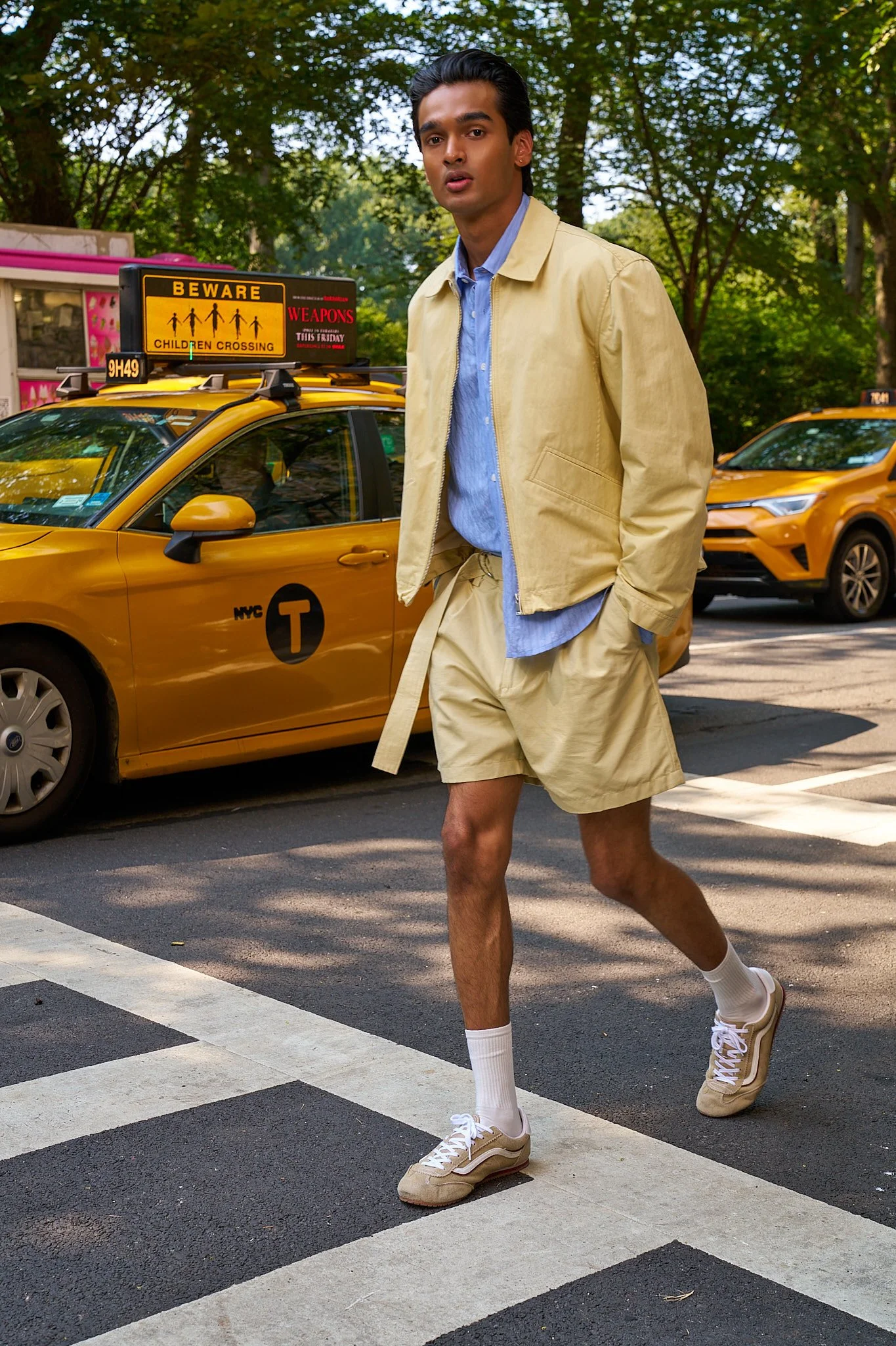 A man in beige shorts and jacket, blue shirt, and white sneakers is crossing the street at a crosswalk. He has dark hair and is looking at the camera. Behind him are yellow taxis and green trees.
