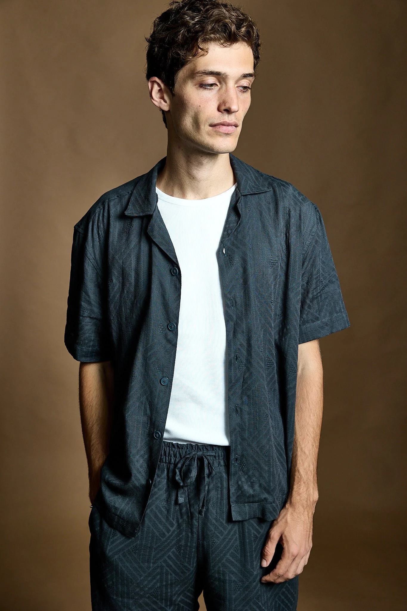 A young man with short, curly brown hair and fair skin stands against a plain brown background. He is wearing a dark, patterned short-sleeve shirt over a white T-shirt and matching patterned shorts. He looks thoughtful with eyes cast downward and his