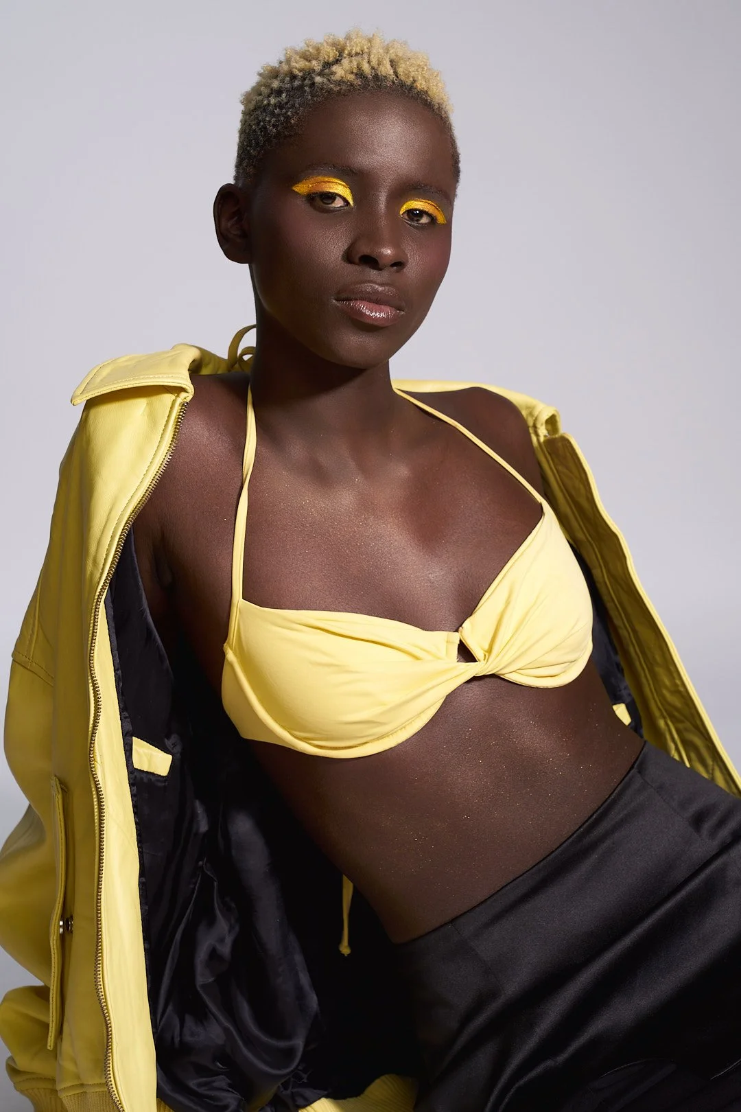 A woman with short blond hair and dark skin wearing yellow eyeshadow, a yellow top, black pants, and a yellow jacket draped over her shoulder, posing against a plain white background.