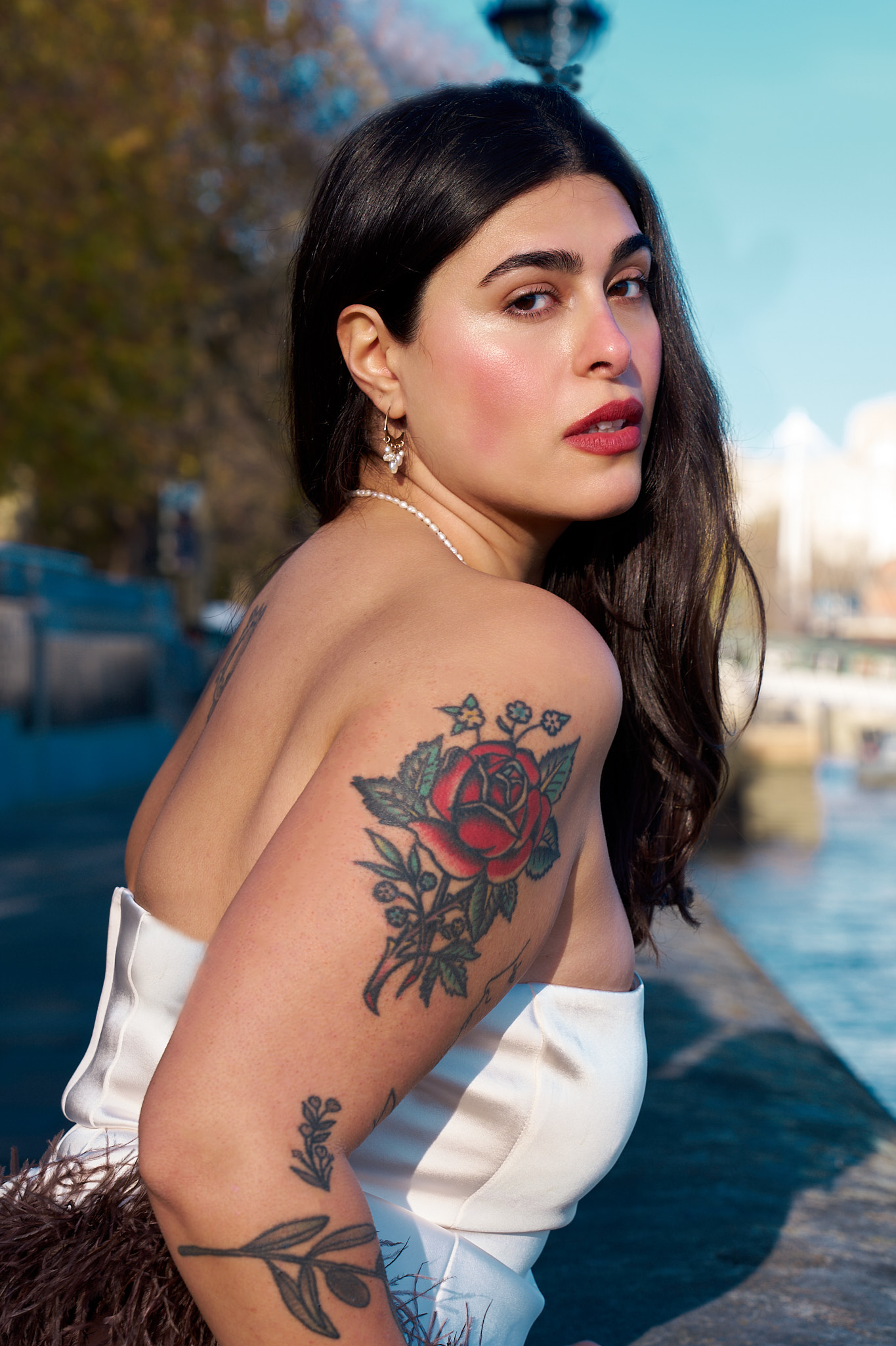 A woman with dark hair and tattoos on her shoulder and arm, wearing earrings, a pearl necklace, and a white top, looking over her shoulder near water with trees and buildings in the background on a sunny day.