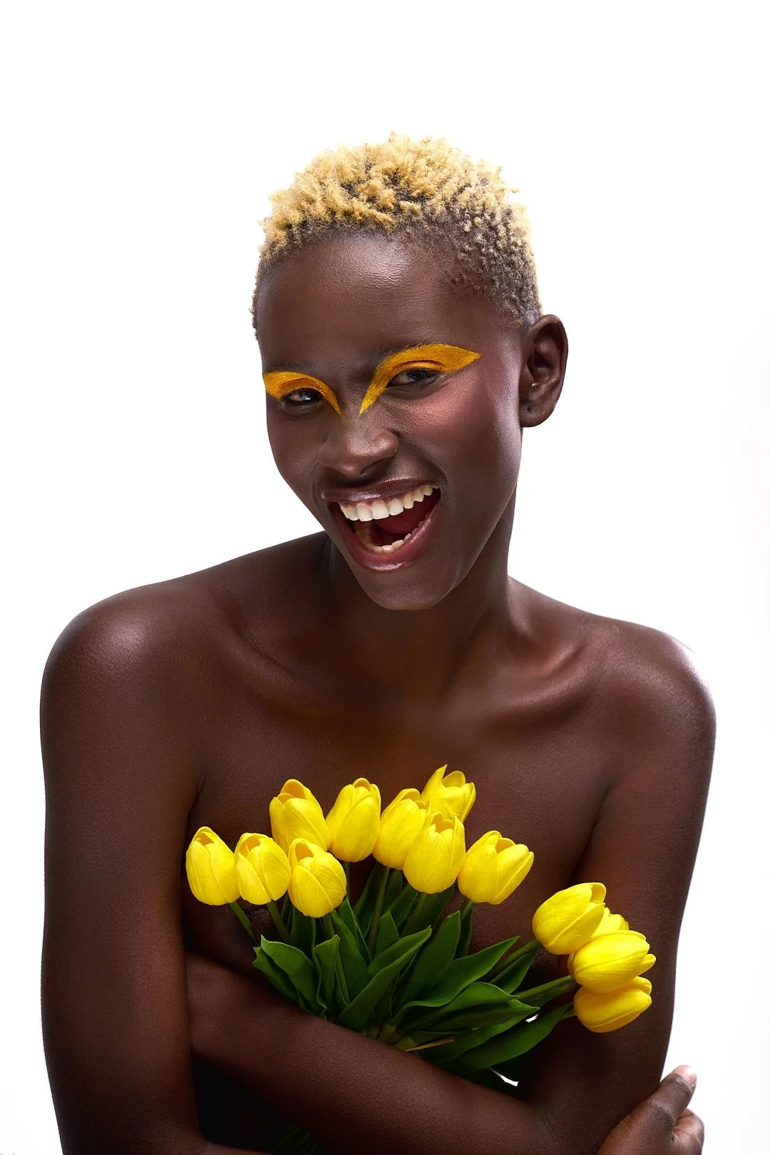 A person with short, blonde, curly hair holding a bouquet of yellow tulips. They are smiling with their mouth open and wearing yellow eye makeup.