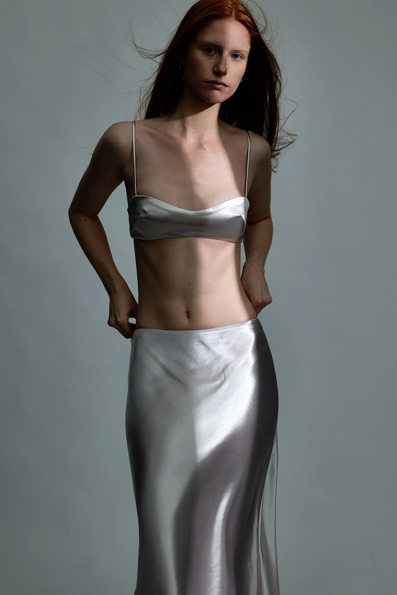A woman with long red hair wearing a silver satin crop top and matching long skirt against a plain gray background.