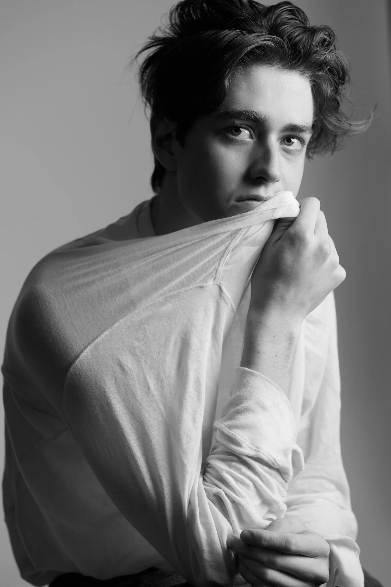 Black and white portrait of a young man with tousled hair, looking directly at the camera with a serious expression, wearing a white shirt with the collar partially covered by a hand.