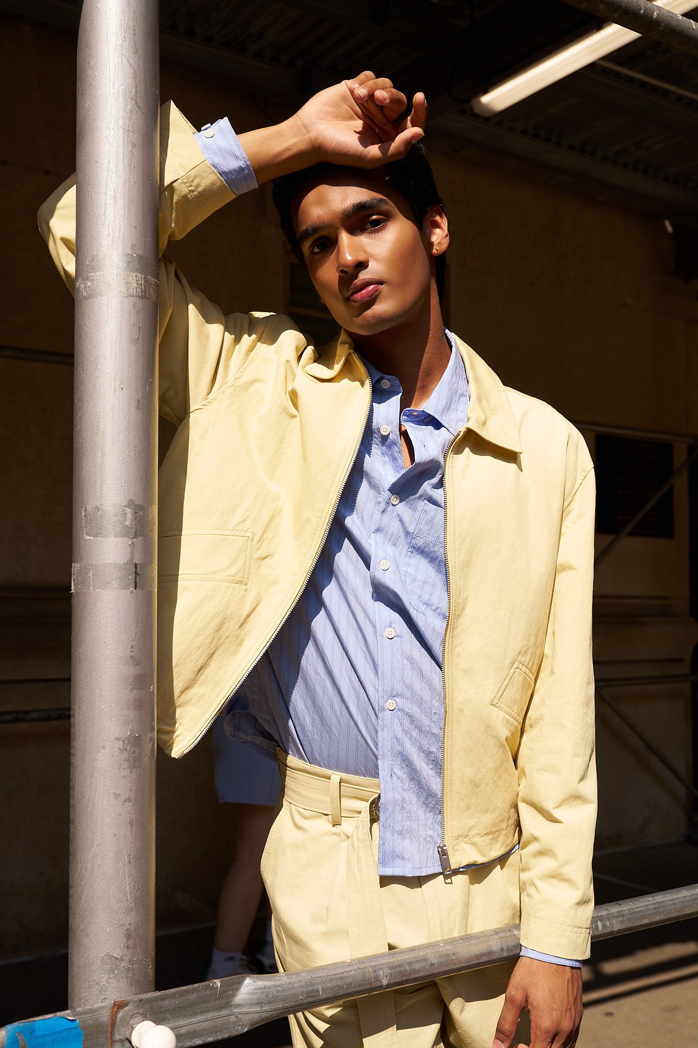 Young man leaning against a metal beam outdoors, wearing a yellow jacket over a blue striped shirt, with a neutral expression and sunlight illuminating his face.